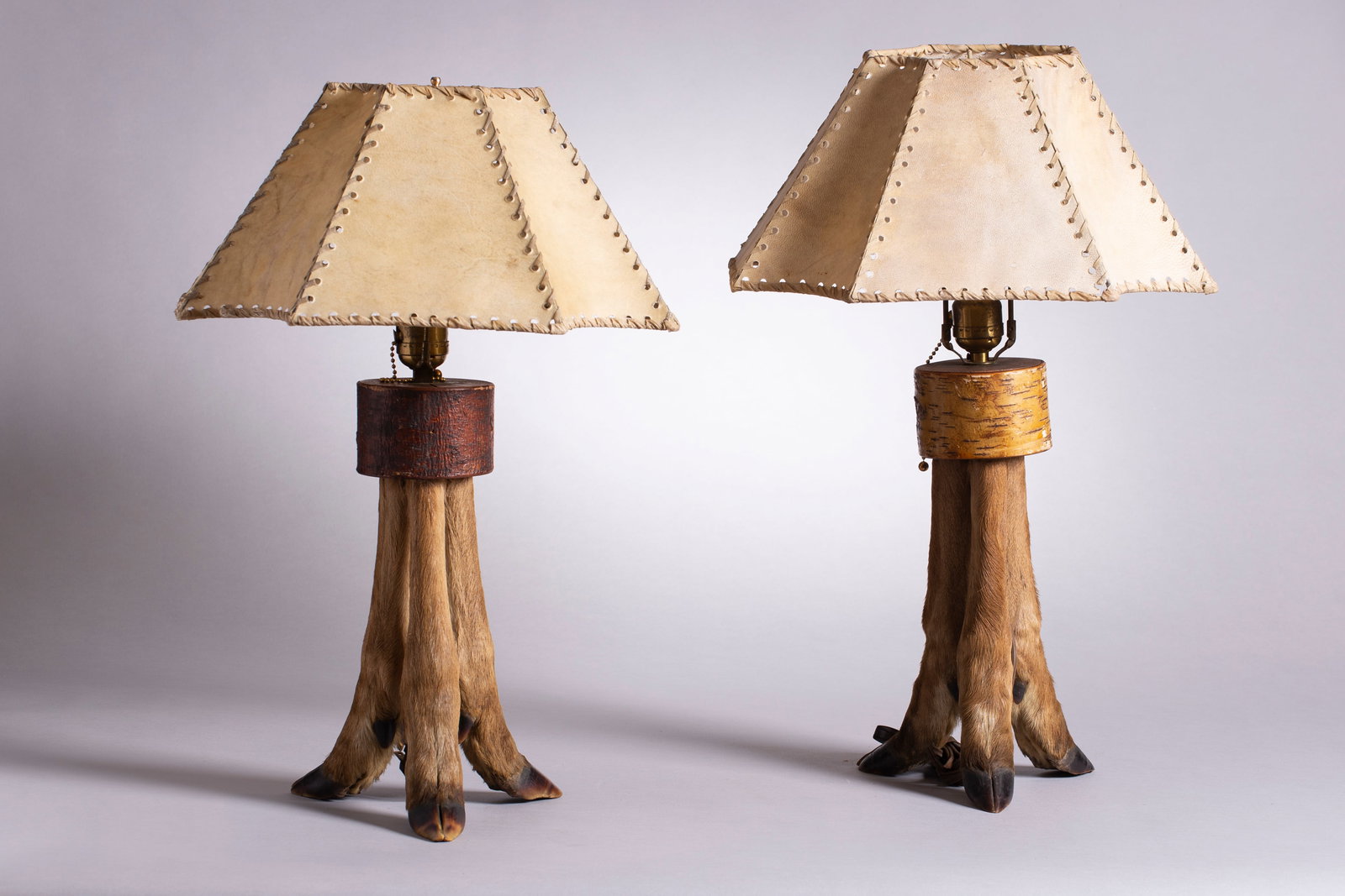 Lot of 2, Deer Hoof Lamps, Ca. 1920s: Lot of 2 American rustic table lamps composed of tri-hoofed deer legs with vintage whipstitched hide shades, Ca. 1920s