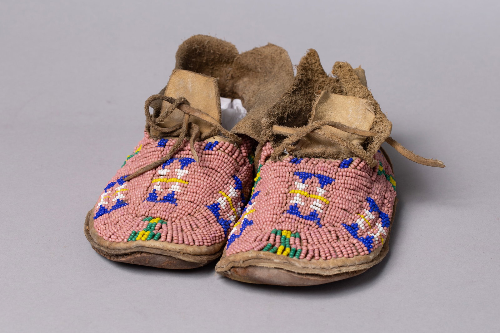 Lot of 2: Pairs of Children's Beaded Moccasins: 1) Soft hide uppers, rawhide soles. The vamps and sides stitched with glass seed beads on a rose-pink ground, showing stacked blue hourglass motifs edged with white and yellow, and green–yellow bead