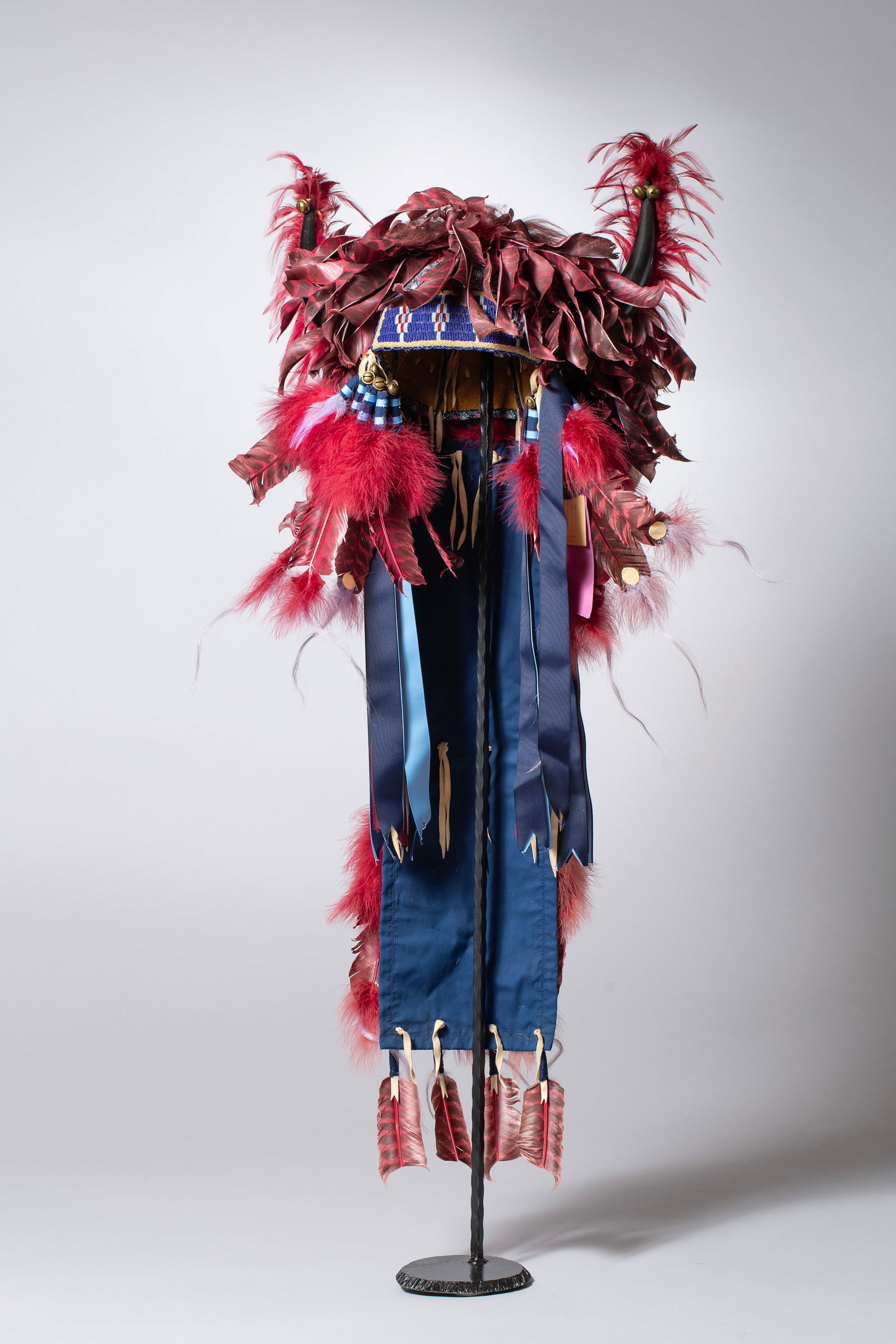 Contemporary Powwow Warbonnet: Award winning elaborate horned and feathered headdress, with split buffalo horns, beadwork, covered with burgundy fluffs and feathers 50" x 21". Does not include stand.