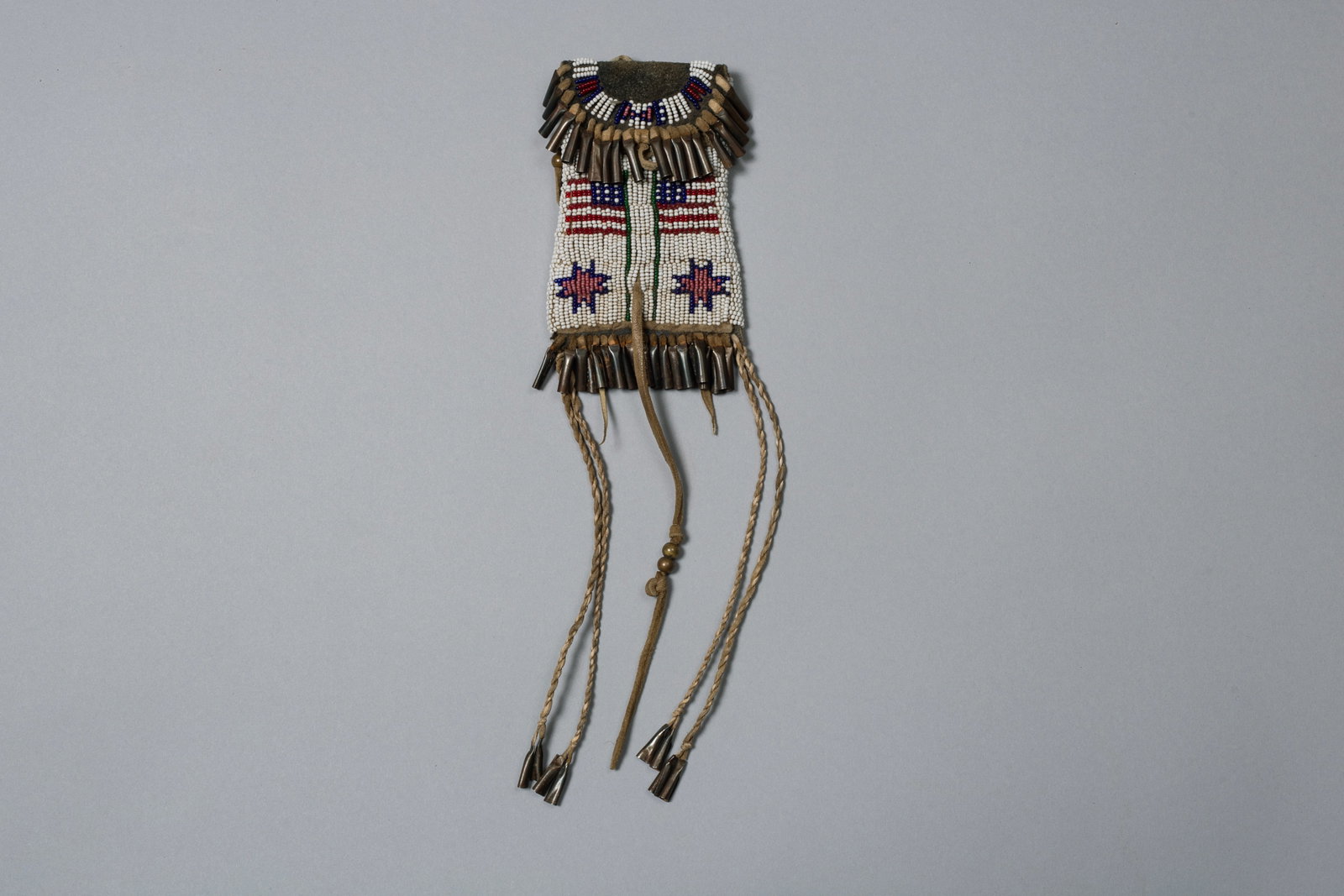 Northern Plains Strike with American Flags: Hide strike-a-light bag beaded on a white ground featuring American flag motifs paired about two eight-pointed star design elements in the lower corners, with tin cones around the flap and at the bott