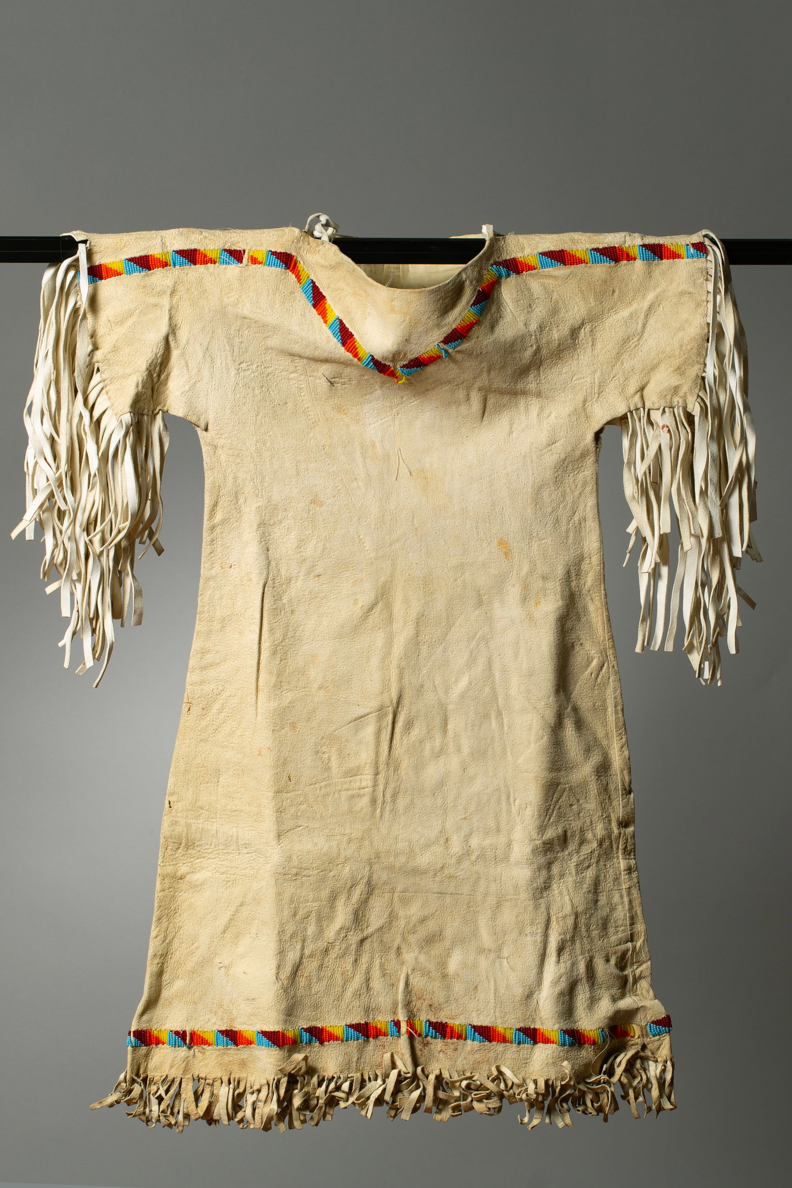 A Plains Beaded Buckskin Girl's Dress: Featuring a traditional, two panel, pattern, the V shaped strip of seed beads of medium blue, dark red, orange, and yellow around the neck and shoulders and around the waist, narrow hide fringe trims 