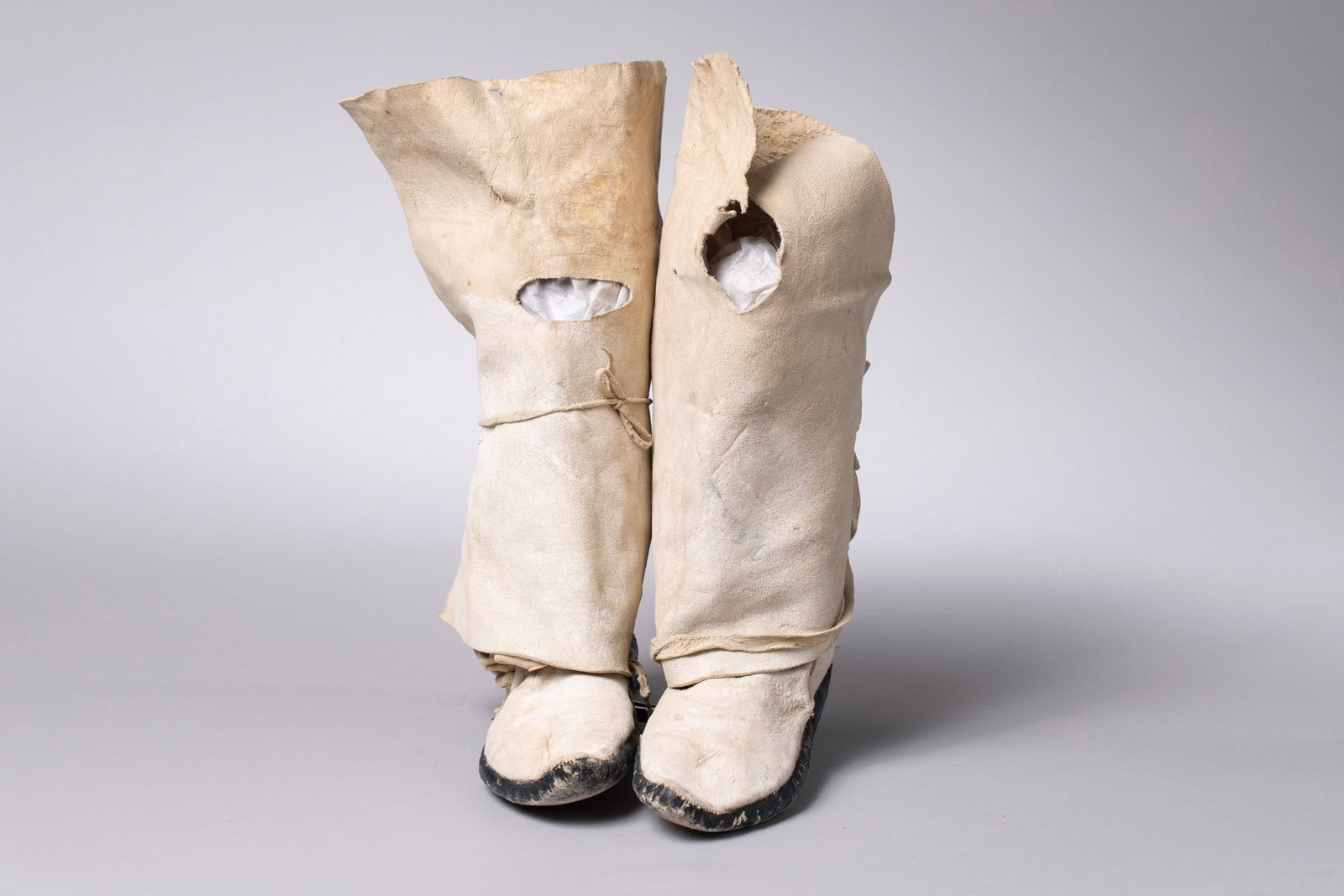 Taos Mocassins: Brain-tanned hide, each moccasin with wraparound high-top leggings, laced with hide ties. The moccasins feature black-painted soles, with soft, rounded toes and natural hide uppers15" high, 10" length