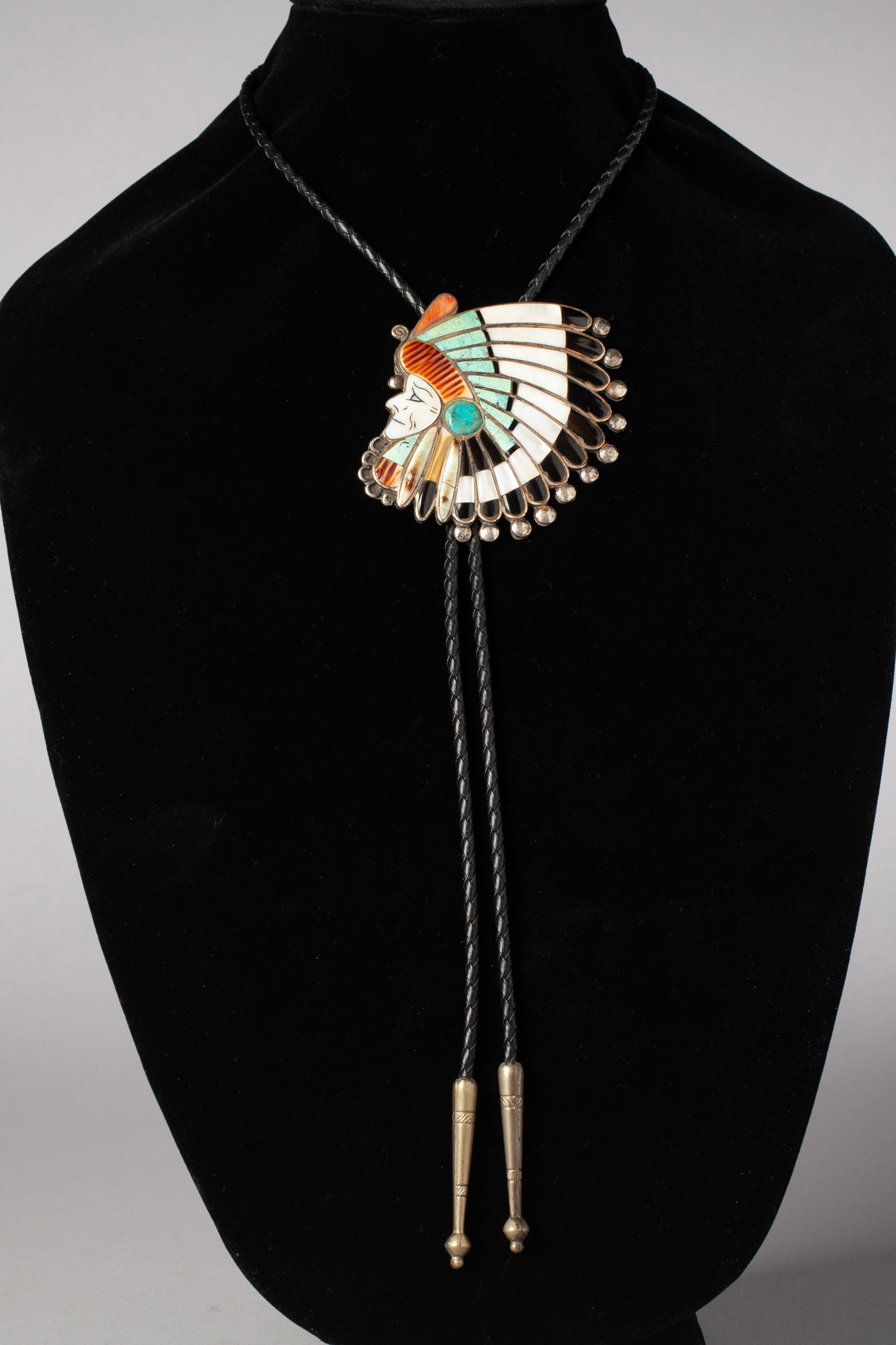 Vintage Zuni Inlay Indian Chief Bolo Tie: Turquoise, mother of pearl, coral, and jet in silver on a black braided leather cord with back clasp 36.5" long including silver cone tips, Bolo measures 3"H, 2 3/4" W