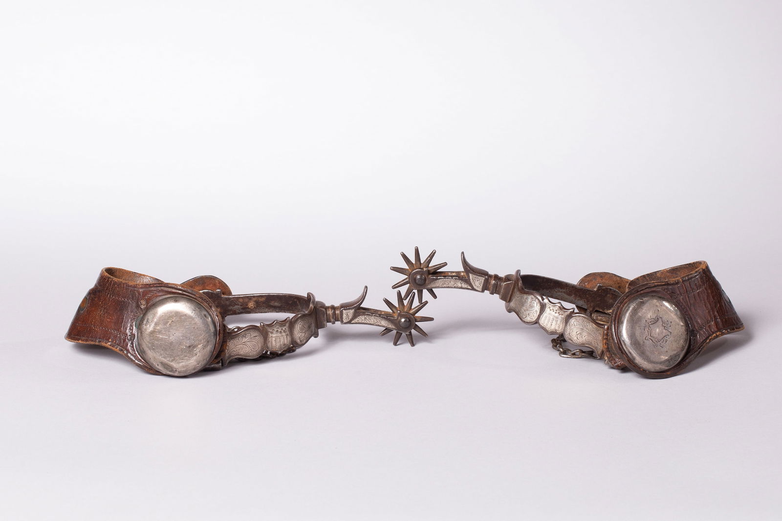 Rare August Buermann Shield Pattern Spurs: Marked with the "Star", silver engraved inlays of the Shield pattern with scroll and the offside has deeply forged pattern in the iron, heel chains, appropriate age straps. Ca. Early 1900s; Heel openi