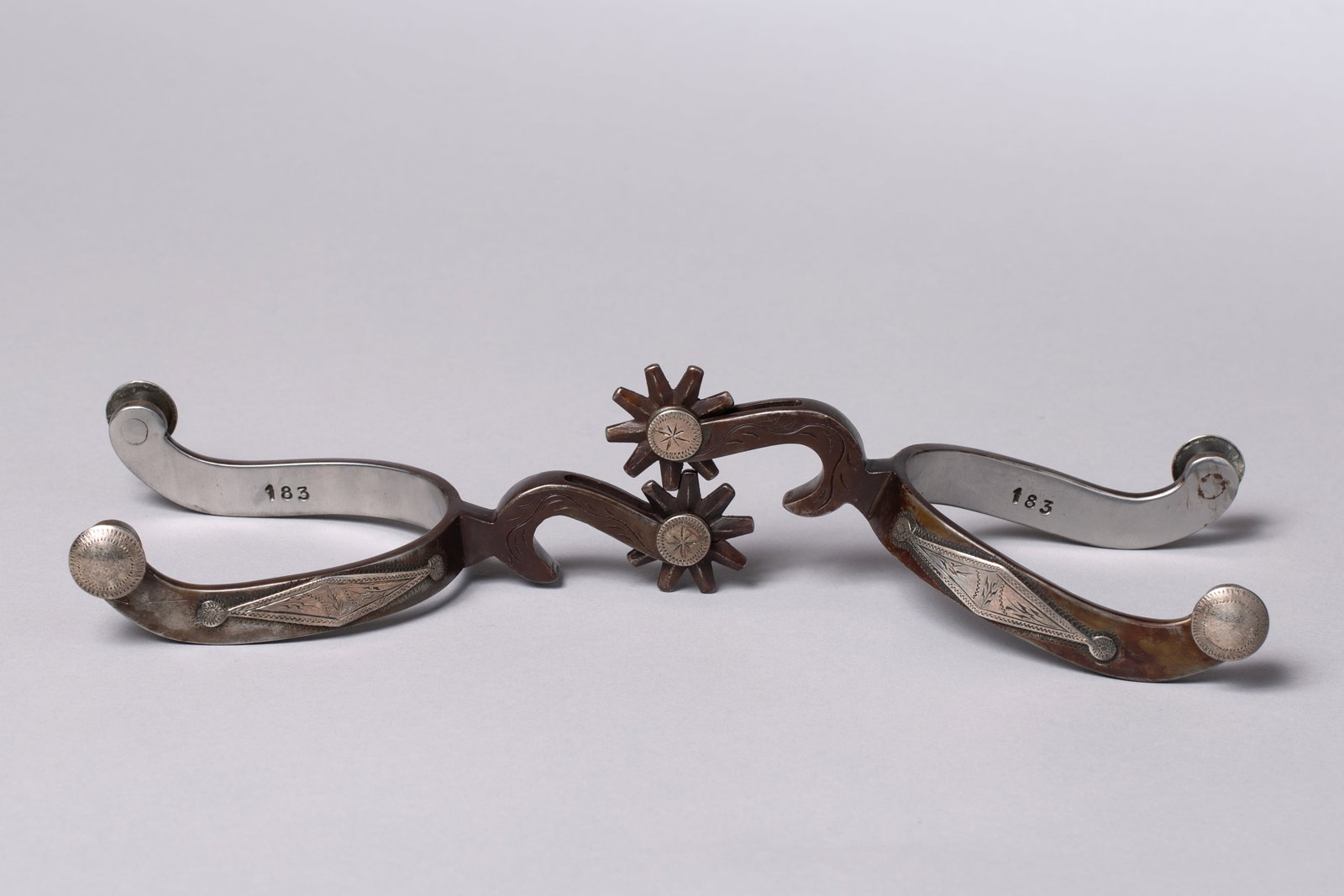 Ron Bliss Bottle Opener Style Spurs: Ron Bliss marked, double mounted, bottle opener style spurs; stamped "BLISS" and "183", silver overlay on heel band and shank; 3 2/10" heel opening, 1/2" heel band width, 2 1/2" shank, 9-point rowel: 