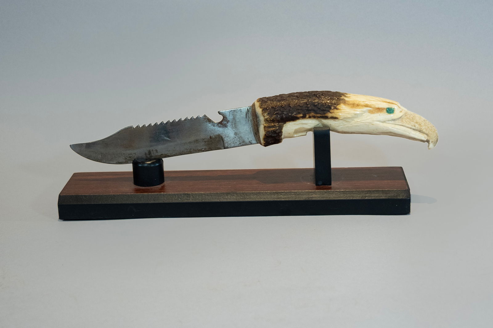 Carved Antler Knife, Ca 1960: Interesting antler handle knife with handle carved into an eagle head. Old blade attached with serrated top and bottle opener. Displayed attractively on included stand. Total length 11 1/4", Blade len