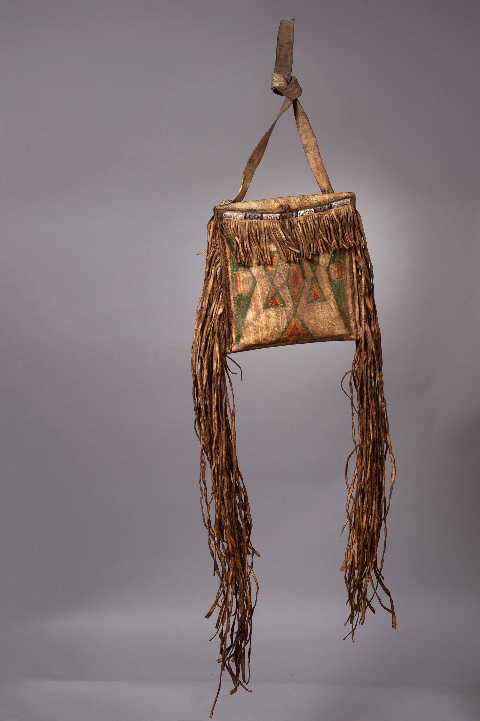 Northern Plains Beaded & Parfleche Bag With Long Fringe: Crafted from sturdy rawhide, this envelope-style bag features striking painted geometric designs in green, red, and yellow earth pigments, a hallmark of Northern Plains artistry. The hand-stitched bea