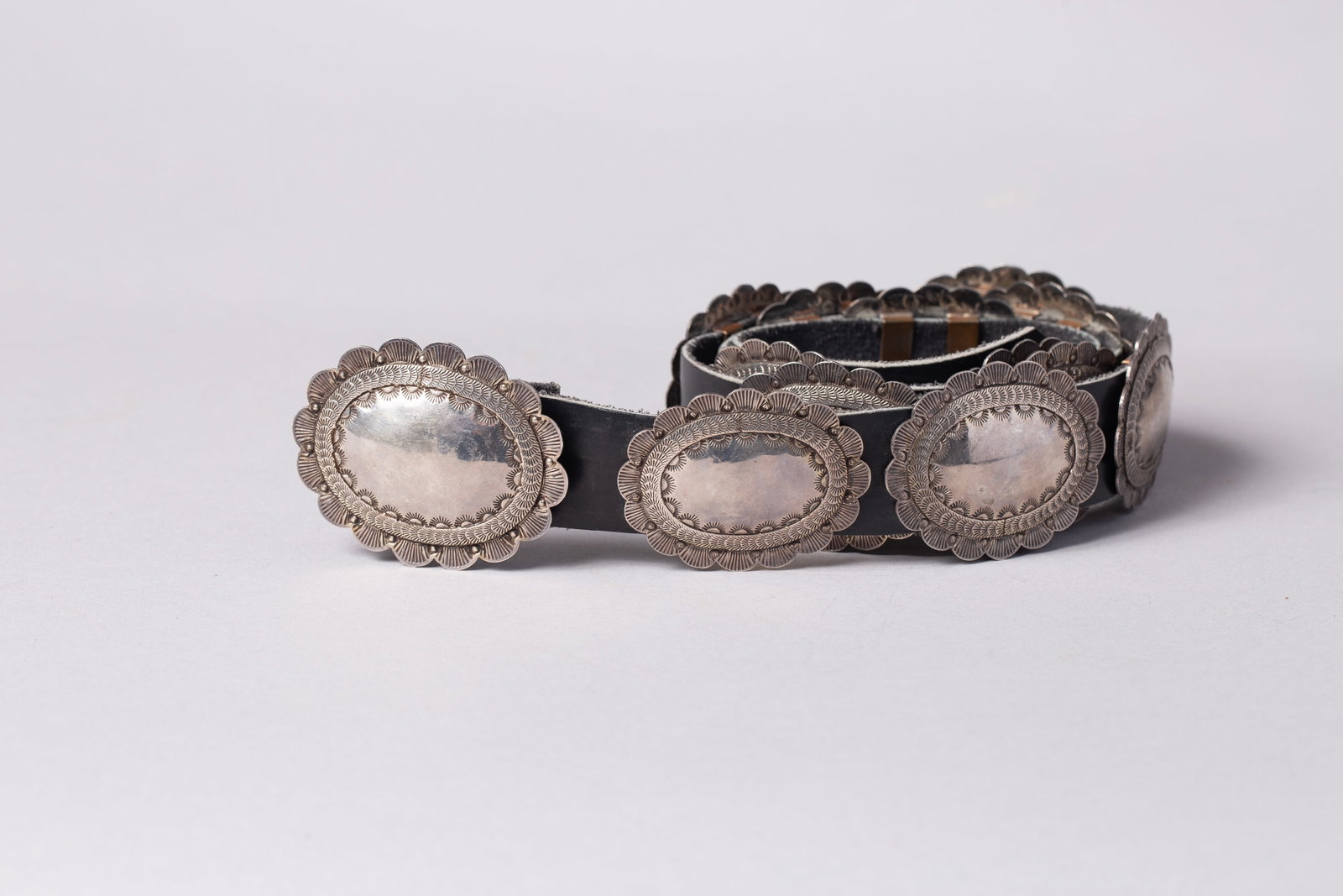 A Native American Stamped Sterling Silver Petite Concho Belt: This finely crafted Southwest sterling silver concho belt features a series of delicate, uniform oval conchos mounted on a dark leather strap. Each concho exhibits classic Navajo-style silversmithing,