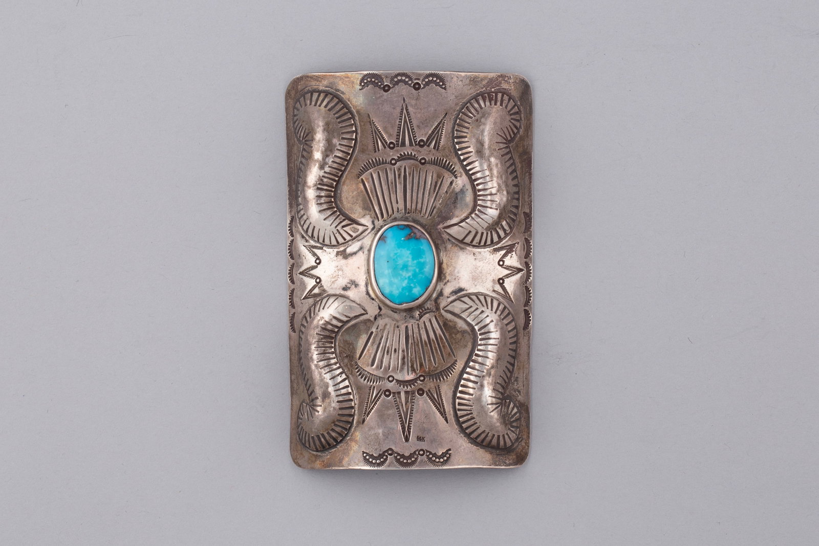 Historic Navajo Ingot Coin Silver Ketoh with Central Turquoise, Ca. Early 1900s: Sterling silver ingot style ketoh featuring a central turquoise cabochon in a smooth bezel setting, surrounded by deeply stamped repoussé and geometric decoration with radiating motifs. This rectangu