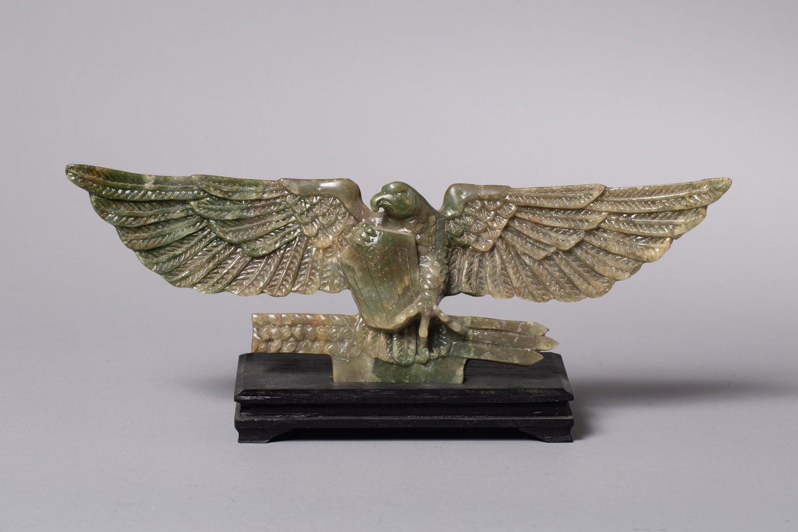 Jade Presentation Eagle: Depicting a spread-wing eagle grasping arrows and shield above a stepped base. Executed in dark green stone with finely carved feather detailing throughout the wings and body, mounted on a wood displa