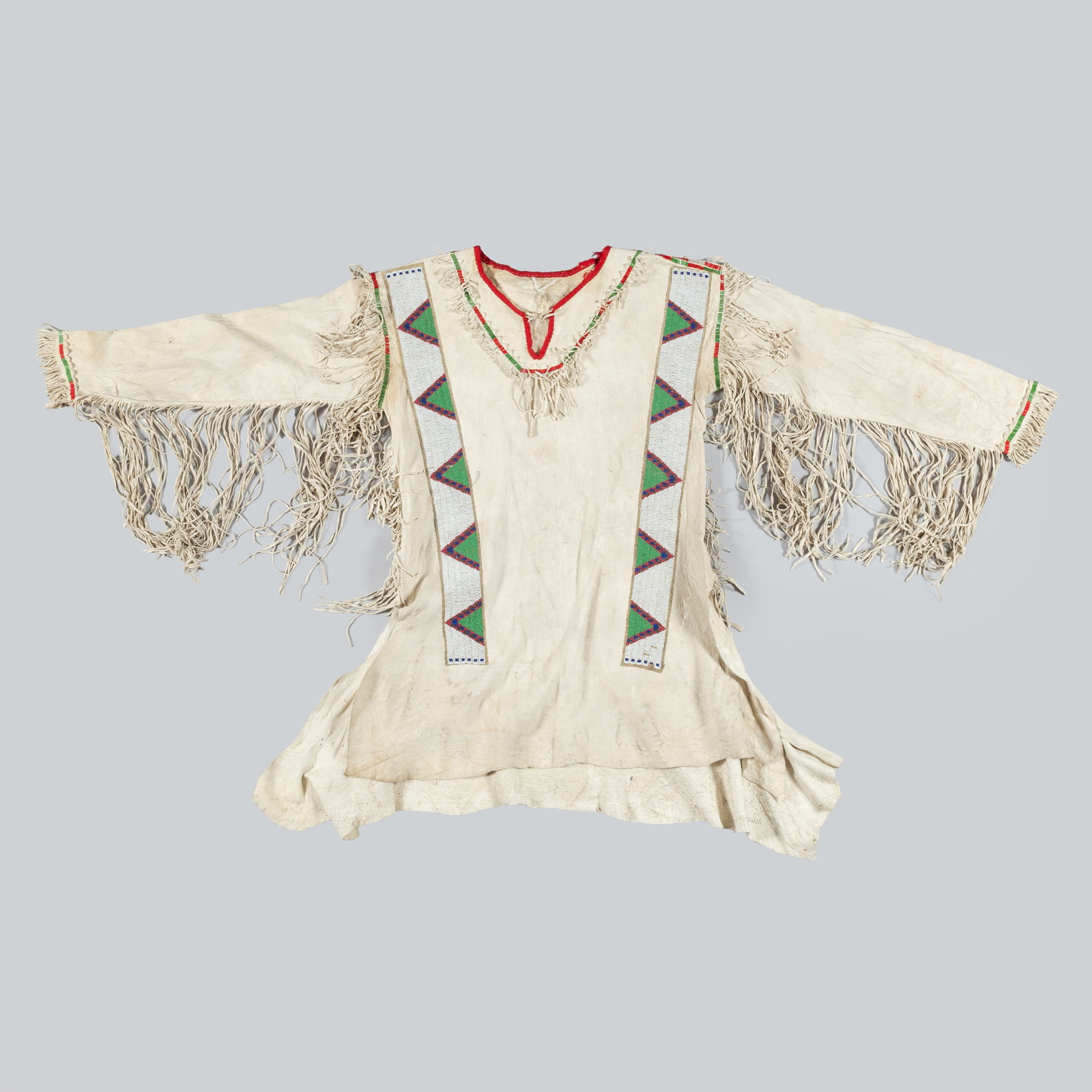 A LAKOTA (SIOUX) BEADED AND FRINGED HIDE MAN’S SHIRT: Lakota shirt constructed of soft tanned hide and adorned with early (1890s) long beaded strips featuring alternating green and purple triangular motifs on a white back ground. The shirt is further emb