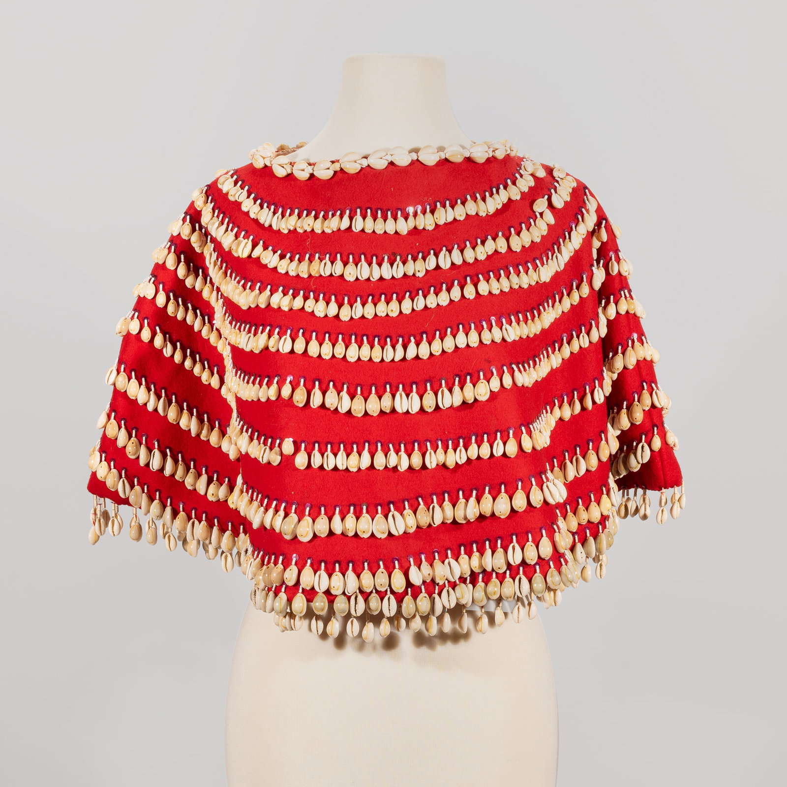 Contemporary Native American Red Shelled Yoke/Cape: A contemporary Native American poncho-style yoke capelet crafted from vibrant red trade cloth. The garment features extensive ornamentation, decorated with numerous concentric rows of stitched cowrie
