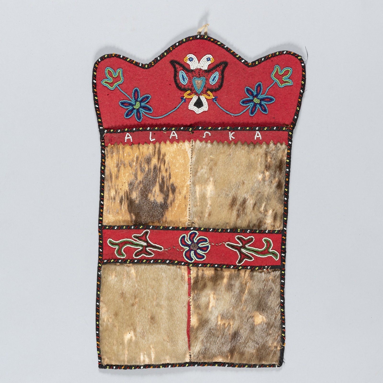 Northwest Coast Beaded Wall Pocket with Seal Hide: Shaped felt top panel in red felt, beaded with a stylized bird motif in white, blue, and outlined detail, flanked by floral beaded motifs in blue and white. Beaded lettering spelling "ALASKA" across