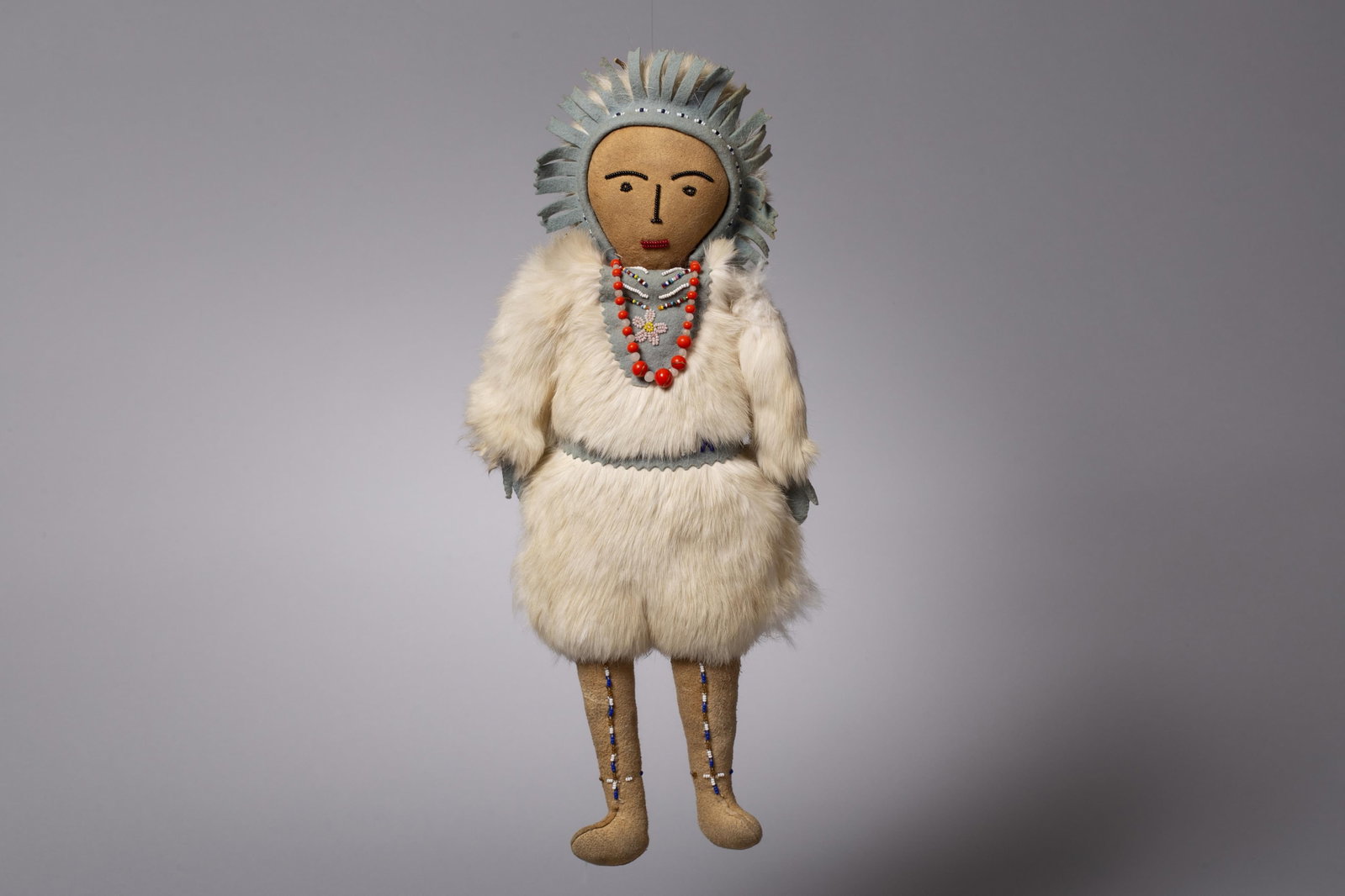Tlingit Style Fur-Clad Doll: A finely crafted indigenous doll featuring a hand-painted wooden face with traditional minimalist features. The figure is clad in a full parka and leggings of thick white Arctic Hare fur, accented