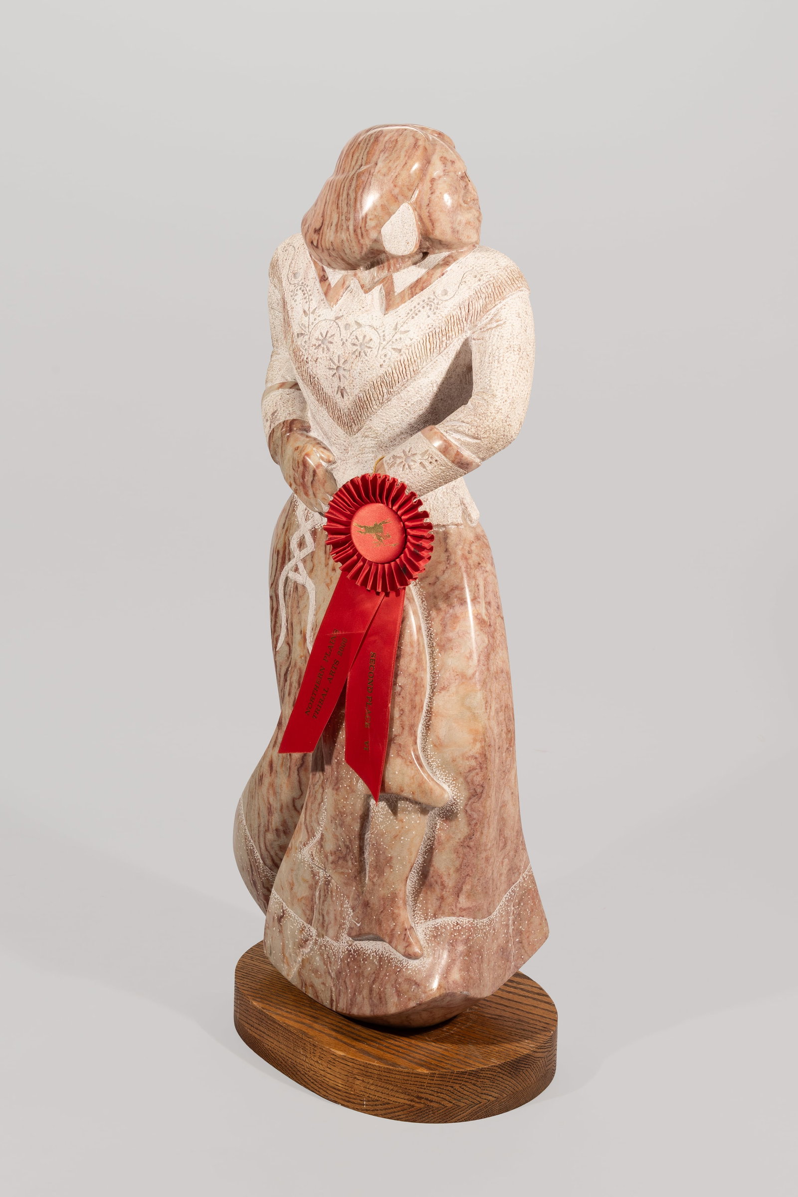 Large Carved Alabaster Sculpture with Northern Plains Tribal Arts 2000 Award: A large and beautifully stylized carved and polished alabaster figure depicting a Native American woman standing with her hands gracefully clasped before her. The figure is wearing a long dress with f