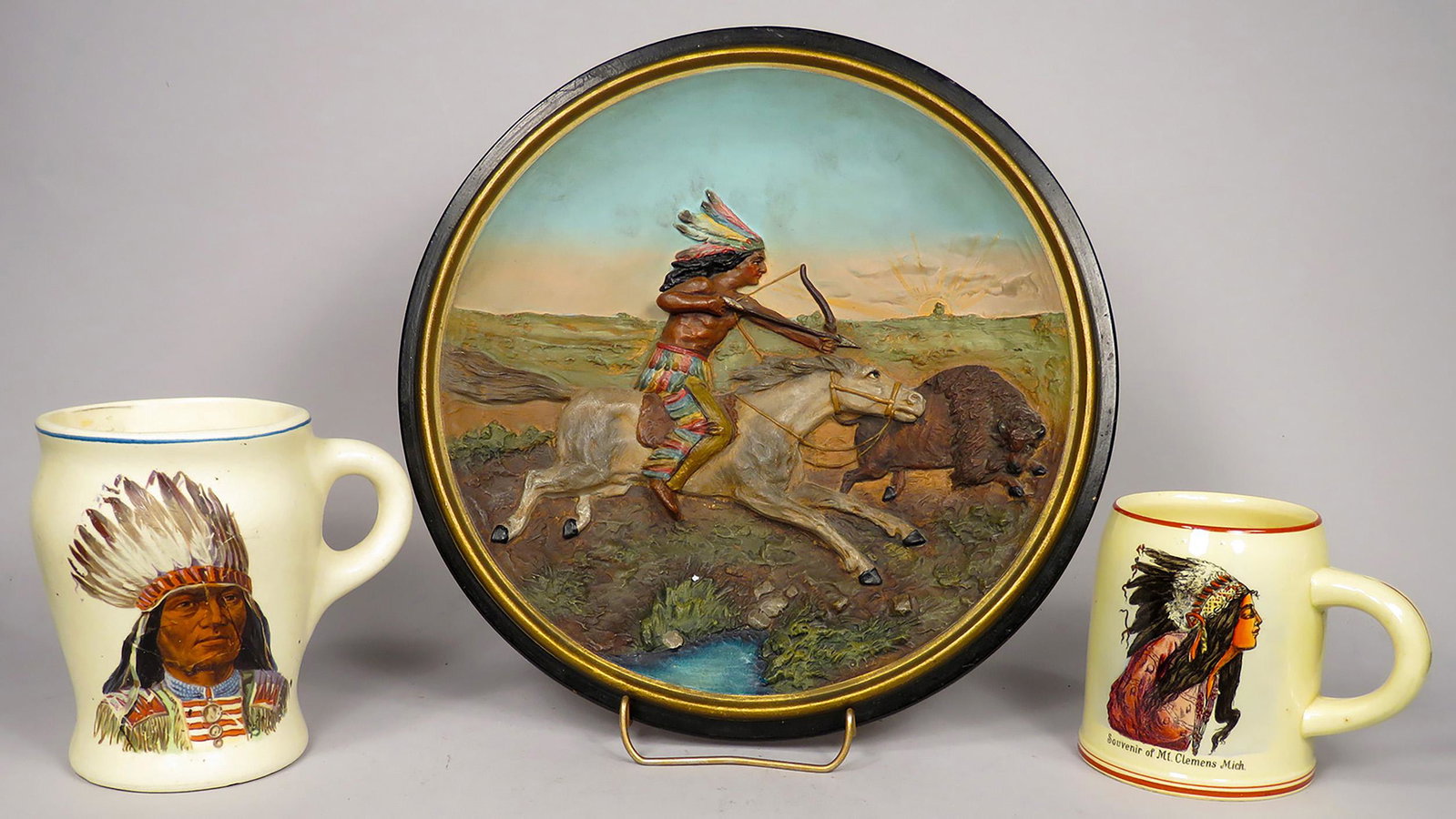 Native American Motif Mugs and Decorative Plate: Native American Motif Mugs and Decorative Plate. Dimensional plate depicting buffalo hunt, stamped JM-5985 on back, measures 10 1/4 inch diameter. Souvenir mug from Mt. Clemens Mich., Made in Germany.
