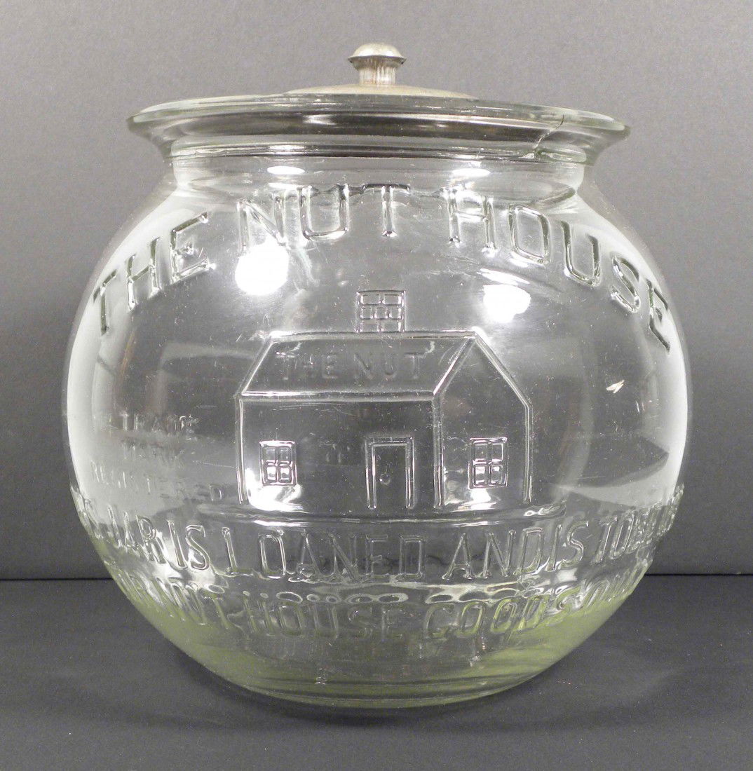 "The Nut House" Embossed Glass Store Jar: The Nut House Embossed Glass Store Jar w/tin lid. "This is loaned and is to be used for Nut House cookies only". A 6 inch crack across the bottom otherwise Excellent condition. 9 1/2 inch diameter X 9