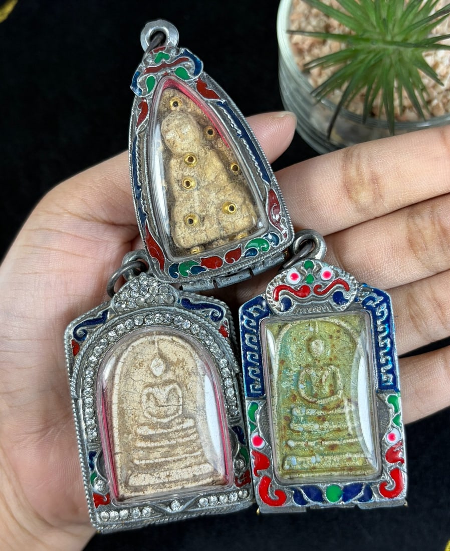 Vintage Thai Enamel Cased Buddha Amulets - 3 Piece Religious Lot, 81AR: This grouping holds three vintage Thai Buddhist votive amulets each housed within untested silvered metal protective casings fitted with clear viewing panels and integrated suspension loops for wear. 