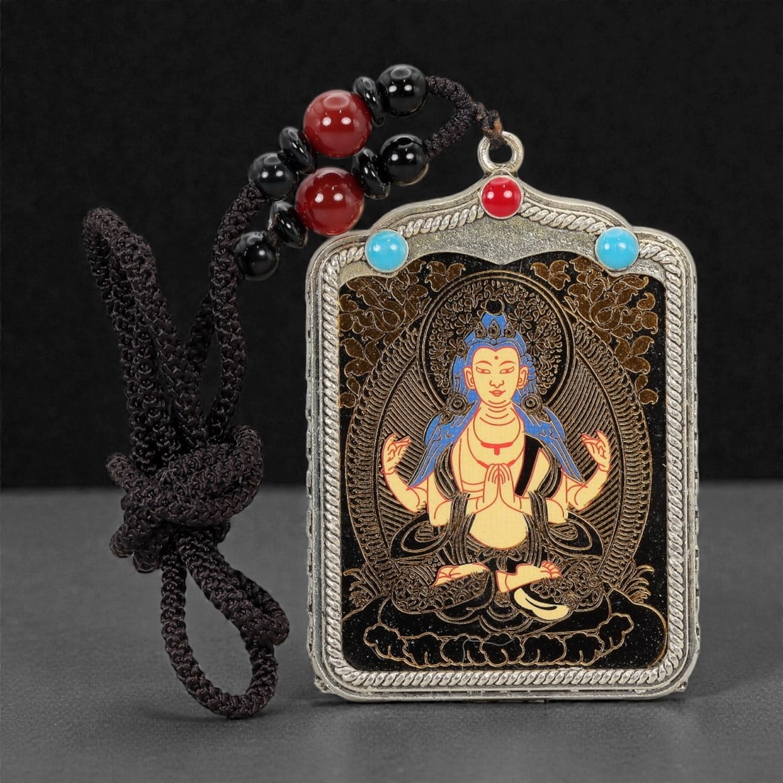 A Tibetan Metal and Painted Amulet, 20th Century, 17FH: The amulet is constructed with a rectangular metal frame housing a painted devotional image, suspended from a braided cord with applied beads. The central panel depicts a seated Buddhist figure shown 