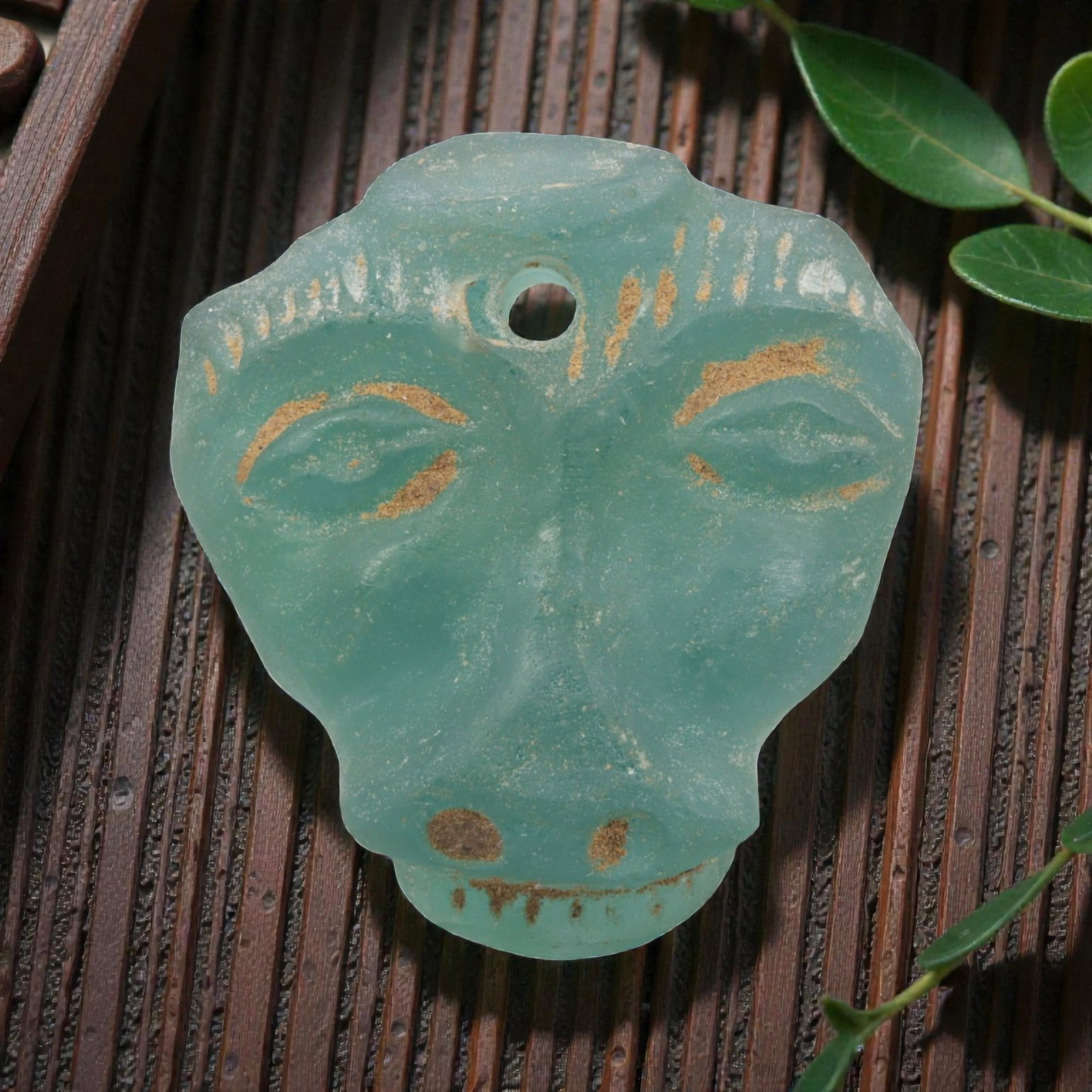 An Ancient Roman Glass Face Amulet Pendant, 9AH: A single ancient-style glass amulet pendant, crafted from translucent turquoise Roman glass, measuring approximately 5 x 4.5 cm. The pendant is carved into a stylized human face with incised eyes, nos