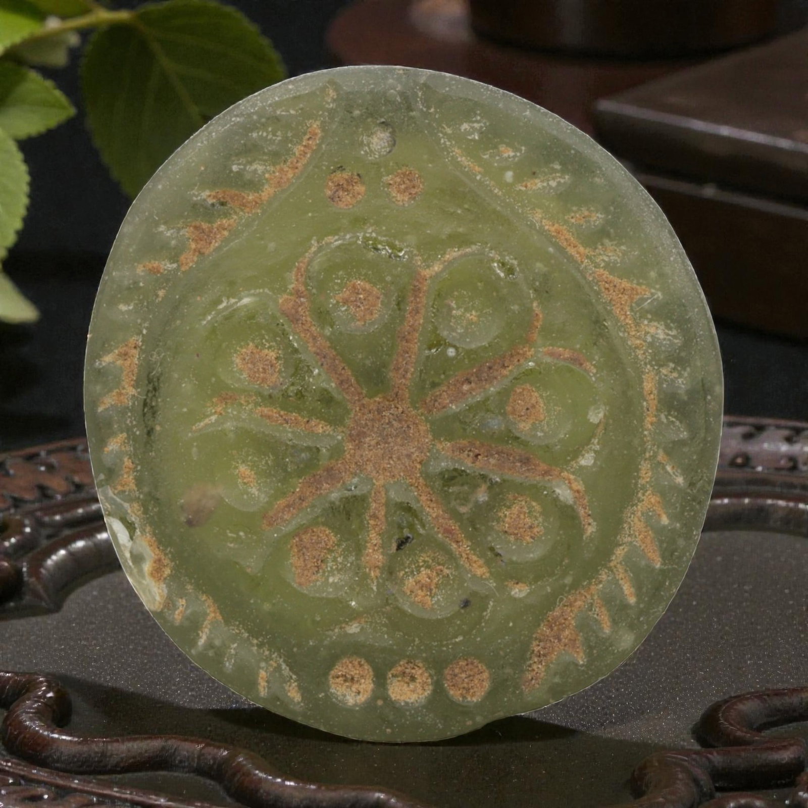 An Ancient Roman Glass Floral Amulet Pendant, 72AH: A single ancient-style glass amulet pendant, crafted from translucent pale green Roman glass, measuring approximately 3.5 x 3 cm. The pendant features an incised floral or rosette motif, with a centra