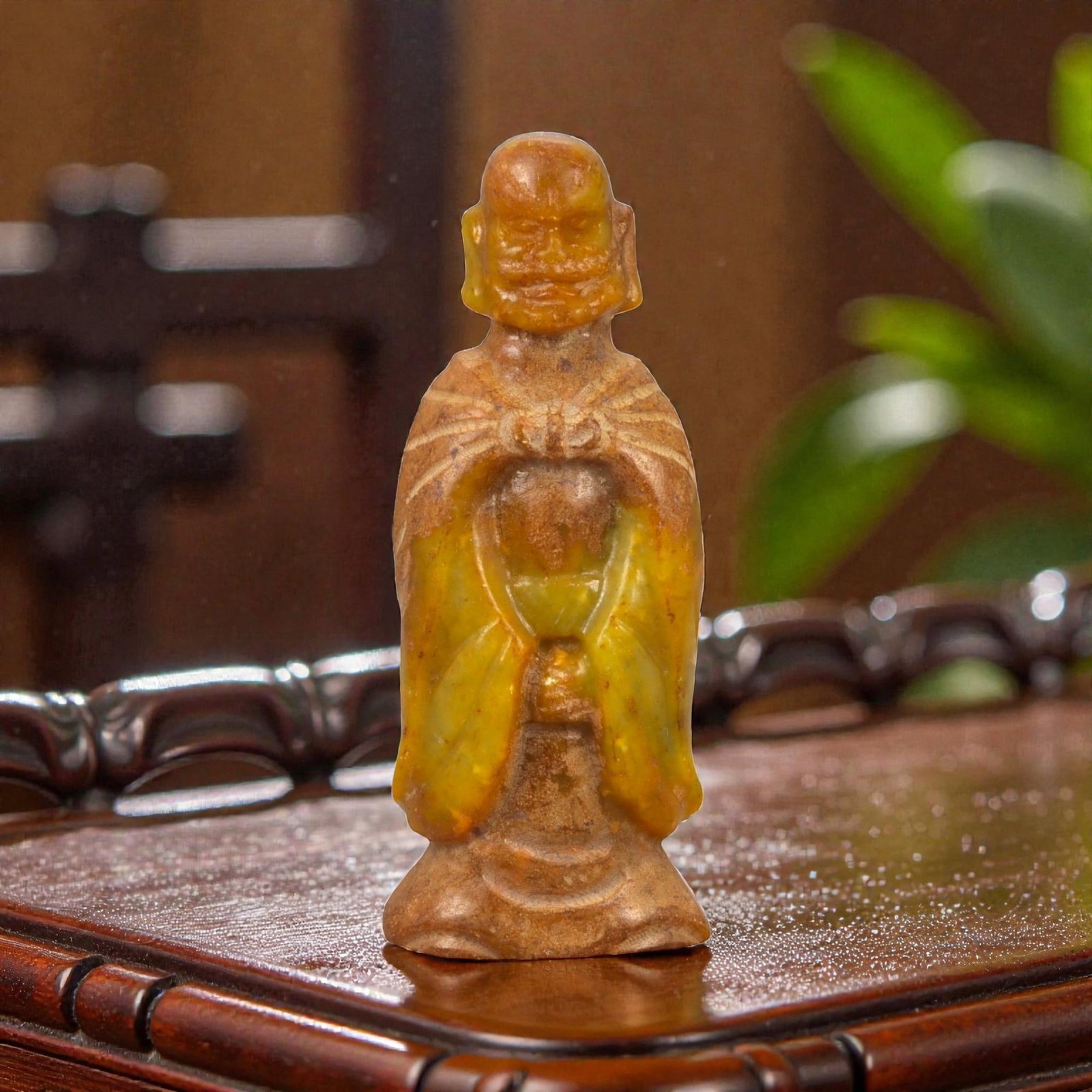 A Chinese Jade Figure of a Standing Monk, 20th Century, 65FB: Carved from translucent yellow to brown-toned jade with areas of darker inclusions and natural surface variation, the figure represents a standing male monk shown frontally. The head is bald, with sim