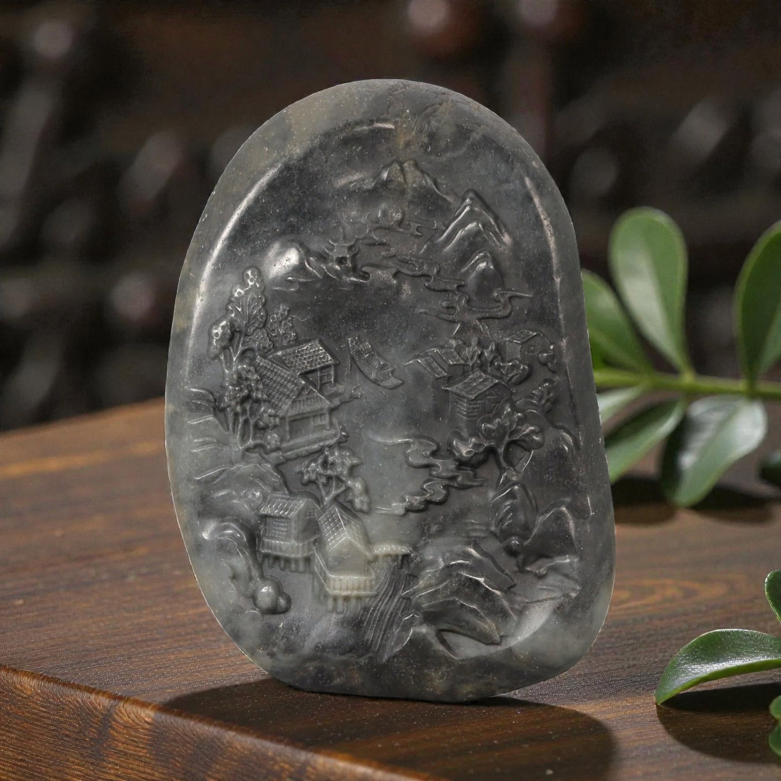 A Chinese Hetian Jade Plaque Carved with a Landscape Scene, 21FD: Carved from dark grey to charcoal-toned Hetian jade with subtle lighter inclusions and natural mottling, the plaque is worked in low to medium relief with a continuous landscape composition. The scene