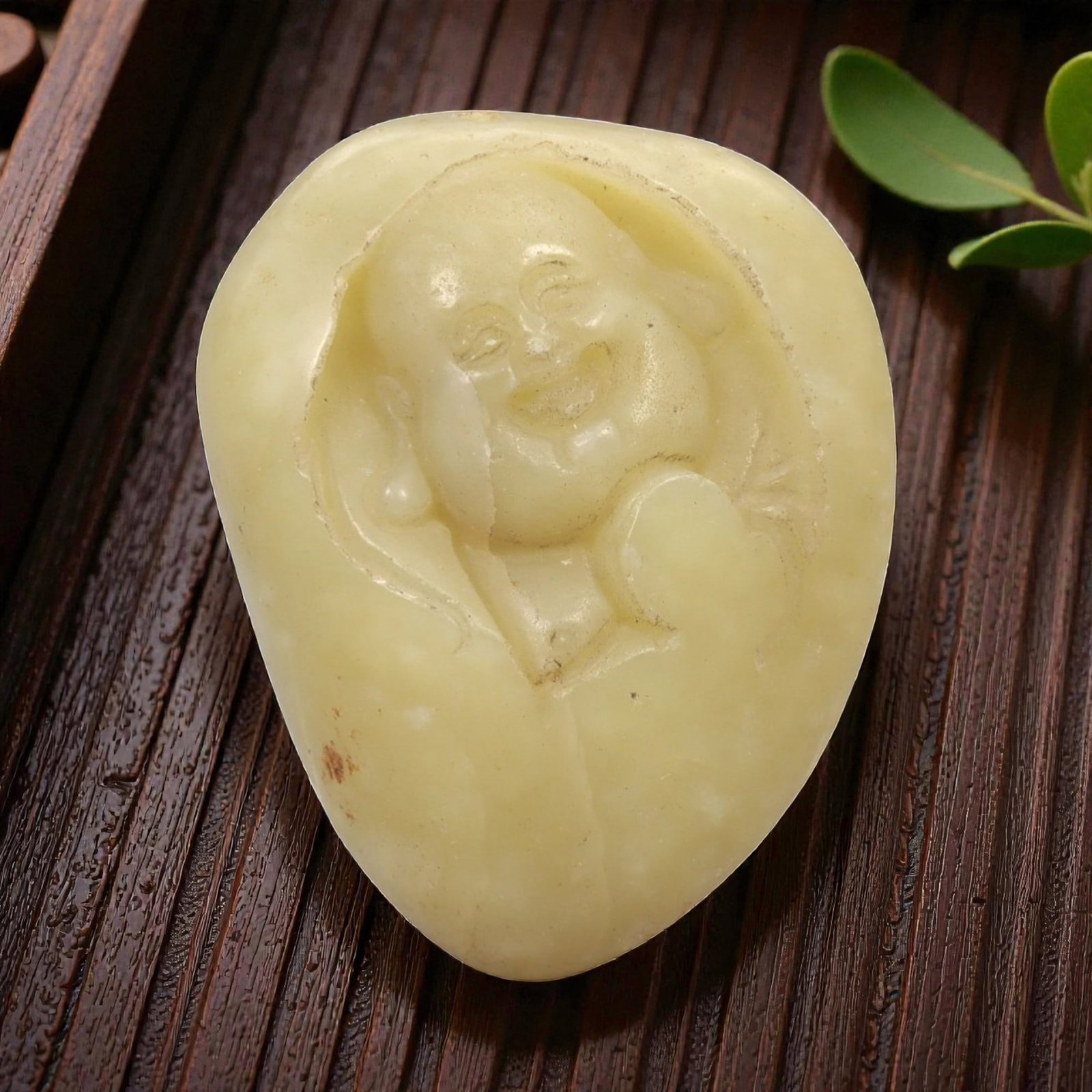 A Chinese Hetian Jade Pebble Carving of Budai, 20th Century, 92FH: Carved from a naturally rounded Hetian nephrite pebble of pale yellowish celadon tone, the carving depicts Budai shown in relief emerging from the natural contours of the stone. The figure is rendered
