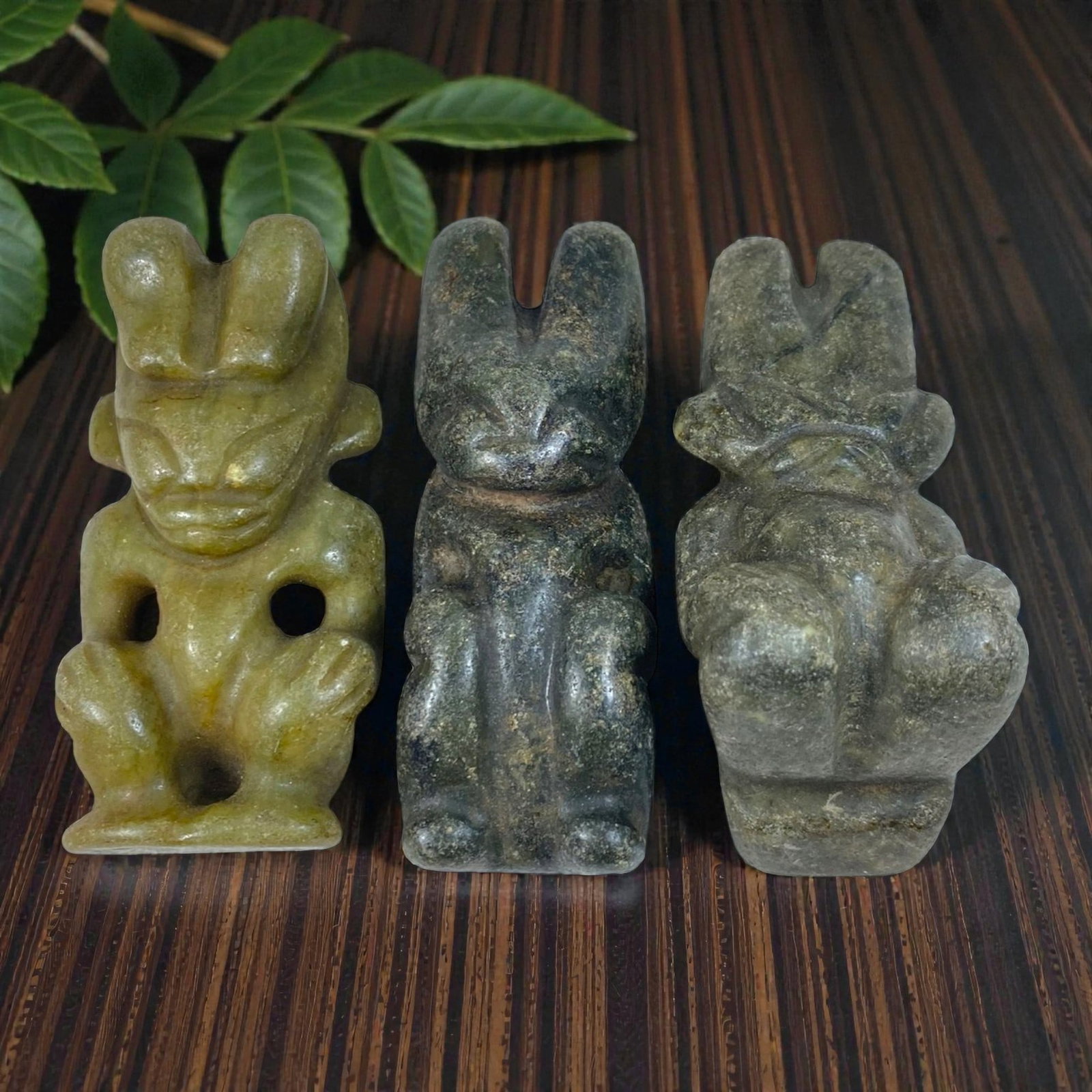 A Lot of 3 Chinese Hongshan-Style Jade Deity Figurines, 65AB: This collection features three Hongshan-style jade carvings depicting seated anthropomorphic deities, crafted from natural jade in varying tones of light green, dark green, and grey-green, exhibiting 