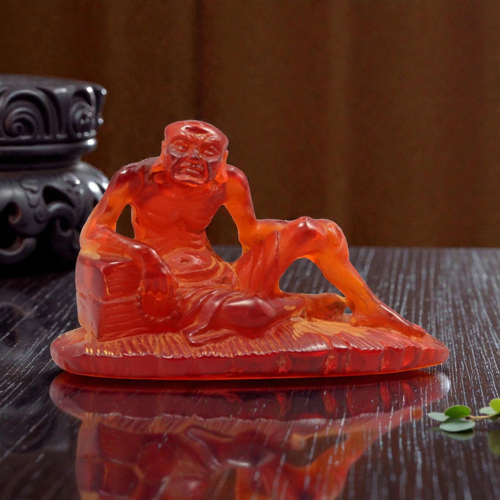 A Chinese Amber-Resin Luohan Figure, 20th Century, 42YC: The figure is molded from translucent amber-colored resin and worked in the round, depicting a reclining luohan shown seated on a low platform. The figure is rendered with an expressive face featuring