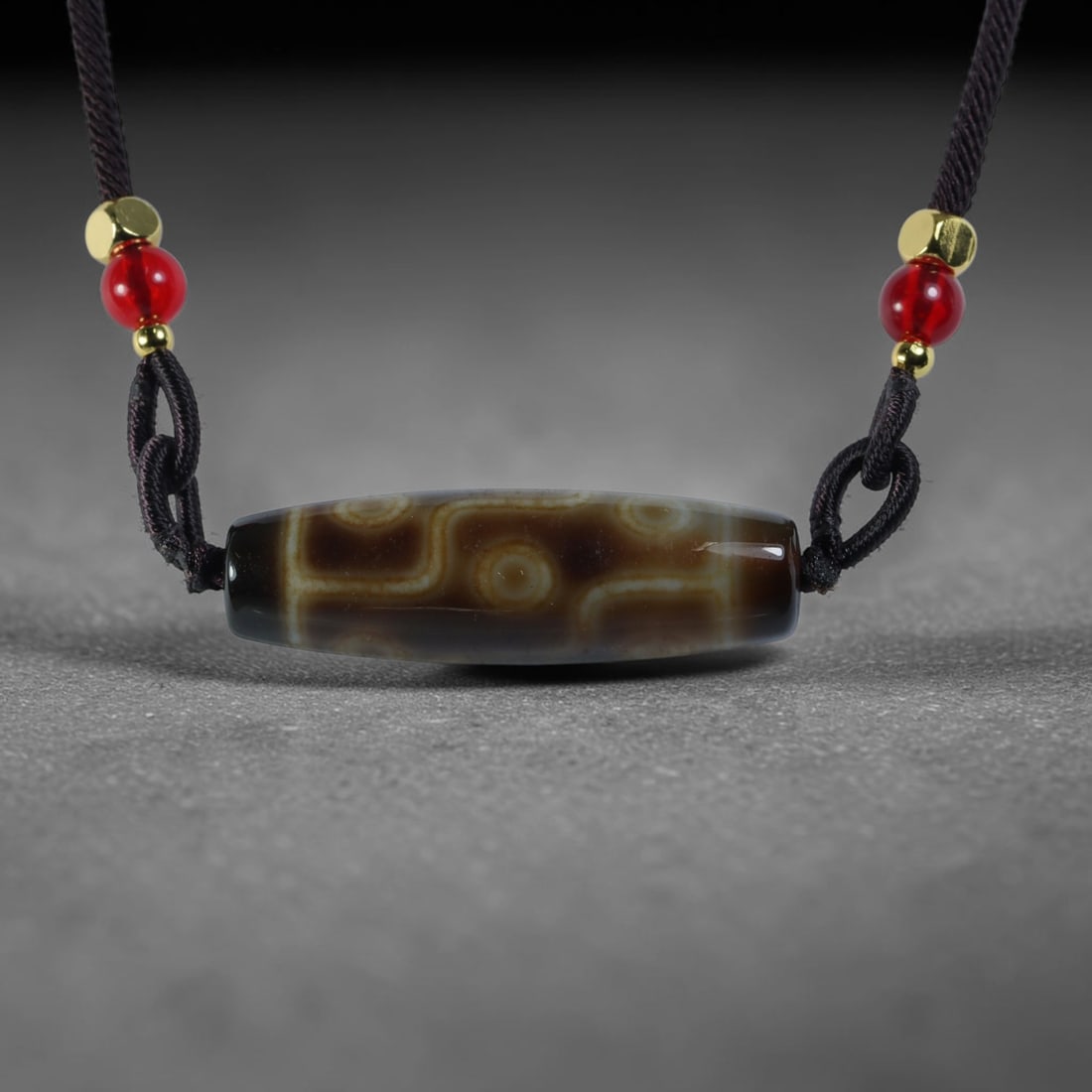 Vintage Tibetan Agate Dzi Bead Strung Pendant, 46YM: Crafted from natural agate with traditional surface patterning, this cylindrical bead has been pre-strung on a woven dark cord fitted with polished red glass accent beads and small metallic spacer bea