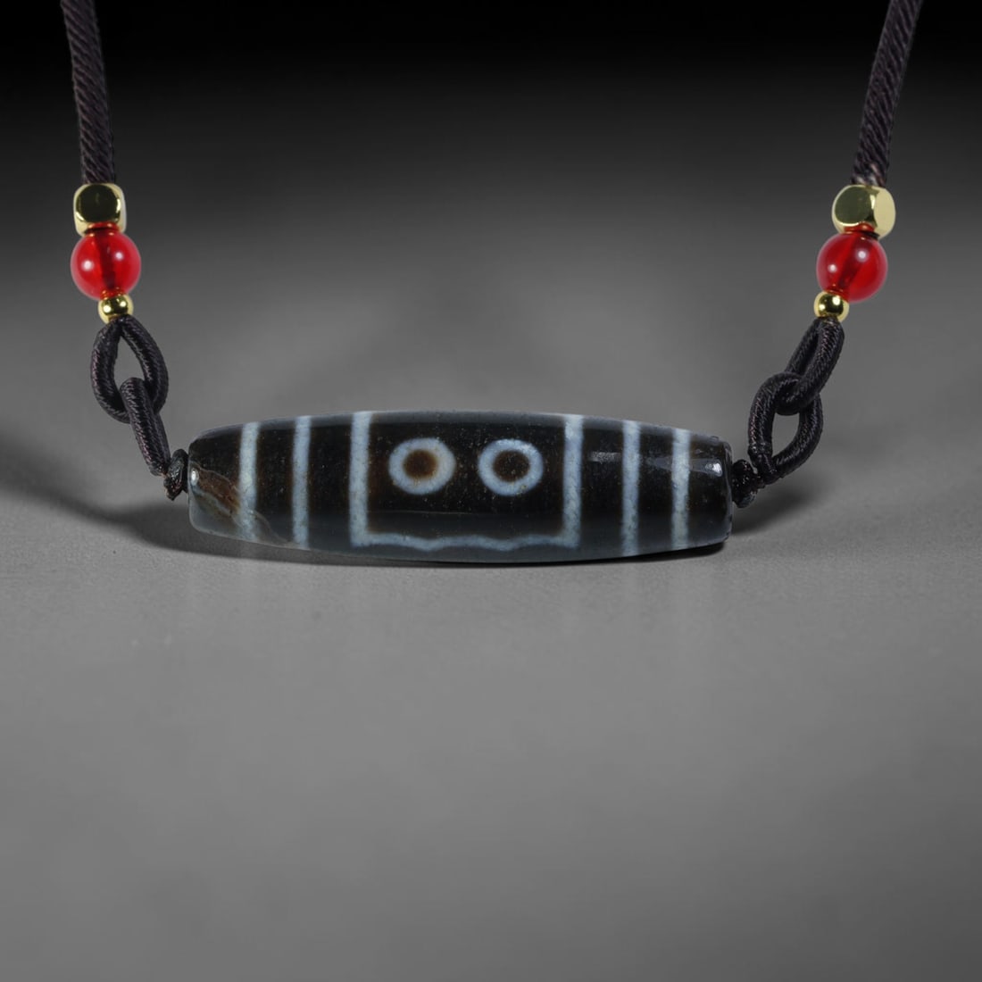 Vintage Tibetan Agate Dzi Bead Strung Pendant, 56YM: Crafted from natural agate with traditional surface patterning, this cylindrical bead has been pre-strung on a woven dark cord fitted with polished red glass accent beads and small metallic spacer bea