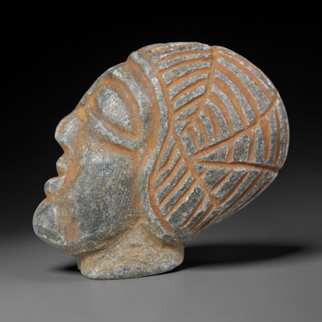 Antique Islamic Carved Stone Head, 36WT: Carved from grey stone with reddish pigment in the incised details, this artifact depicts a stylized human head in profile. The figure features simplified facial features, with a headdress or hairstyl