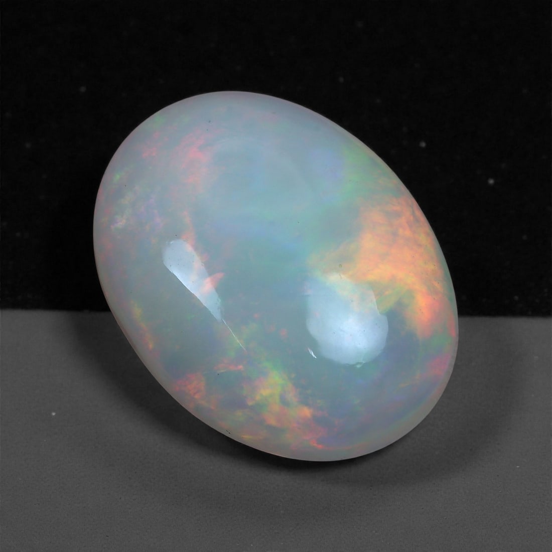 10.46 Carats Greyish Natural Sapphire Cabochon Oval - Natural Ceylon Stone, 18XC: This singular oval cabochon sapphire, measuring 13.8 x 11.6 x 6.1 mm, displays a soft, muted greyish hue with subtle natural variations and a faint warm streak across the surface. The smooth, domed cu