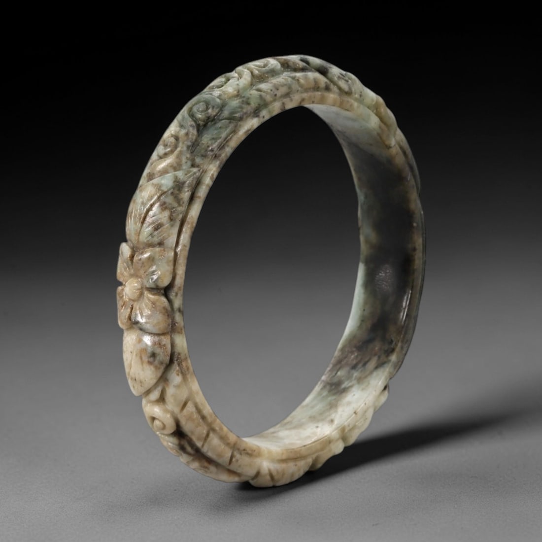 Natural Burmese Type A Carved Jadeite Jade Bangle Bracelet, 55ZL: A finely carved jadeite bangle of flattened circular form, featuring a continuous relief band of scrolling foliate and floral motifs. The stone displays a complex, natural palette of muted celadon-gre