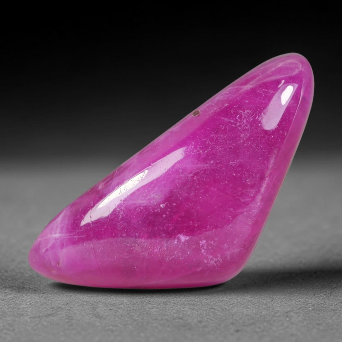 21.79 Carats Natural Madagascar Vivid Fuchsia Pink Heated Ruby Elongated Freeform Cabochon, 89WC: This striking fuchsia pink ruby cabochon displays rich saturated magenta-pink tones covering its smooth polished domed surface, interspersed with faint natural crystalline veining and tiny scattered m