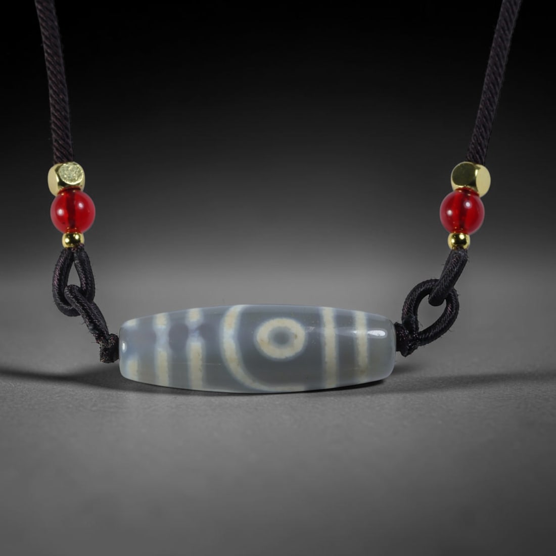Vintage Tibetan Agate Dzi Bead Strung Pendant, 49YM: Crafted from natural agate with traditional surface patterning, this cylindrical bead has been pre-strung on a woven dark cord fitted with polished red glass accent beads and small metallic spacer bea
