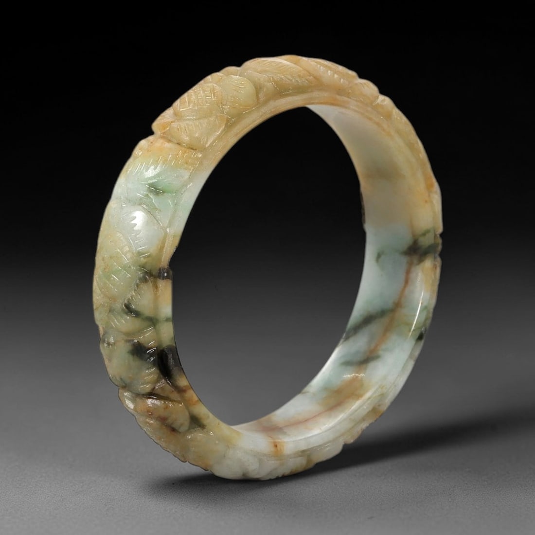 Natural Burmese Type A Carved Jadeite Jade Bangle Bracelet, 39ZL: A petite jadeite bangle of flattened circular form, masterfully carved in relief with a continuous band of scrolling foliate and floral motifs. The stone exhibits a striking variegated palette, with a