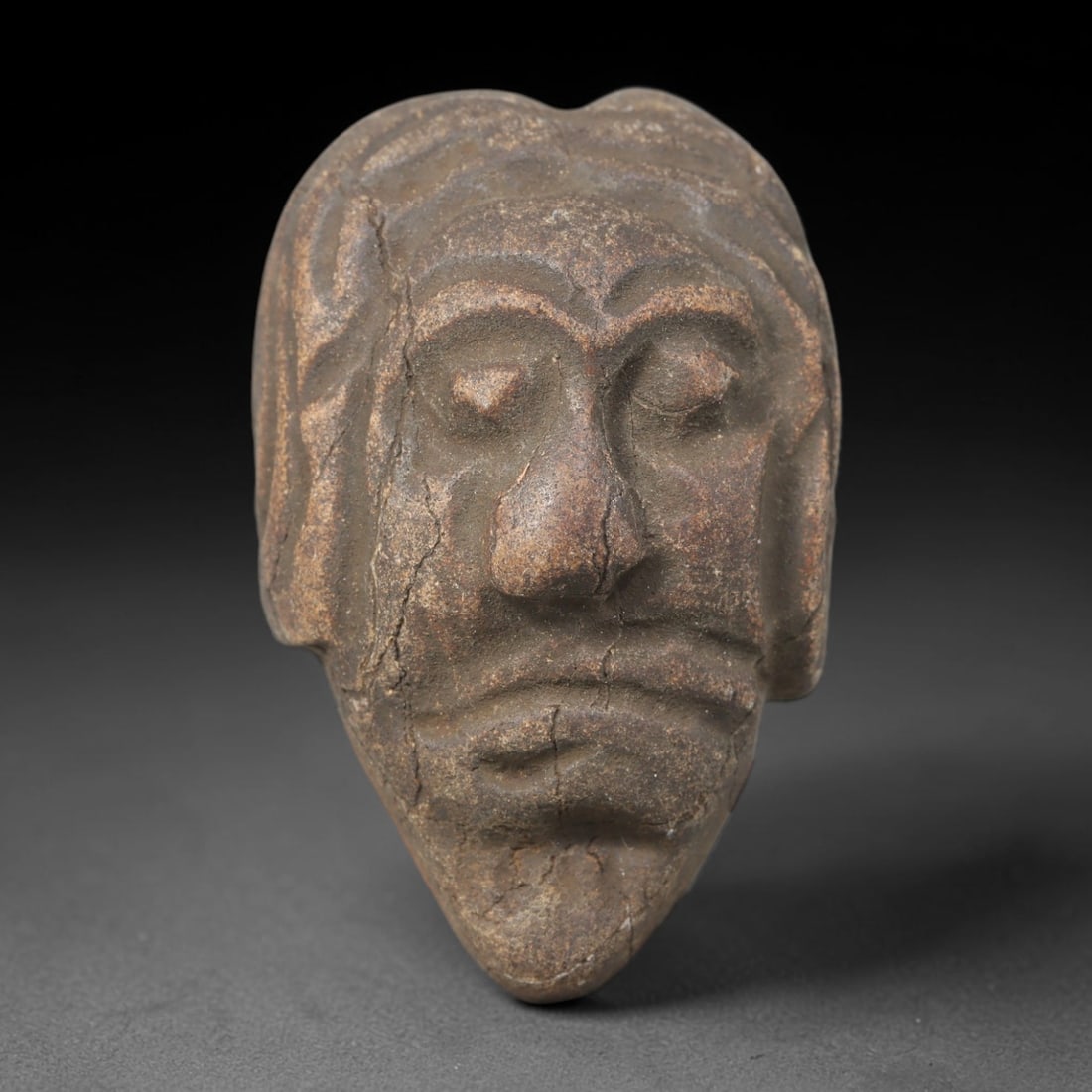 Ancient Medieval Near Eastern Tribal Maskette Stone, 79WT: Crafted from weathered sedimentary stone layered with mottled earthy burial patina, this compact maskette bears hand-carved recessed facial detailing aligned with medieval Near Eastern tribal stonewor