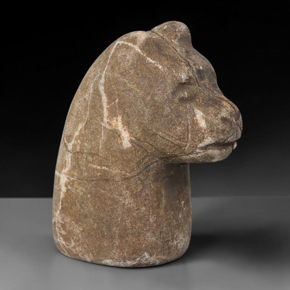 Ancient Medieval Near Eastern Tribal Stone Effigy, 88WT: Sculpted from brown weathered stone threaded with pale mineral veining, this tribal effigy forms a stylized feline animal head mounted upon a solid tapered stone base, with subtle recessed carving def
