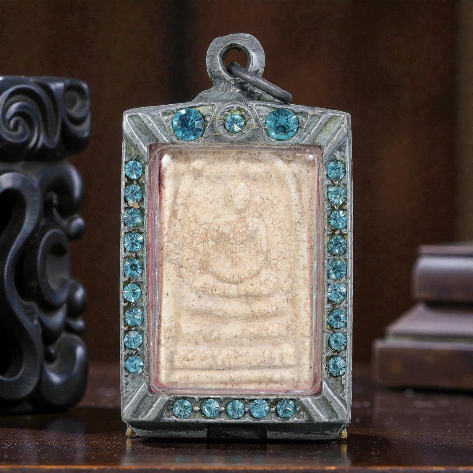 Thai Phra Somdej Amulet - Blue Crystal Silver Tone Pendant, 62ZB: A Thai Buddhist Phra Somdej amulet featuring a seated Buddha figure on tiered bases, housed in a silver-tone metal casing with blue crystal detailing. The glass-fronted enclosure protects the aged amu