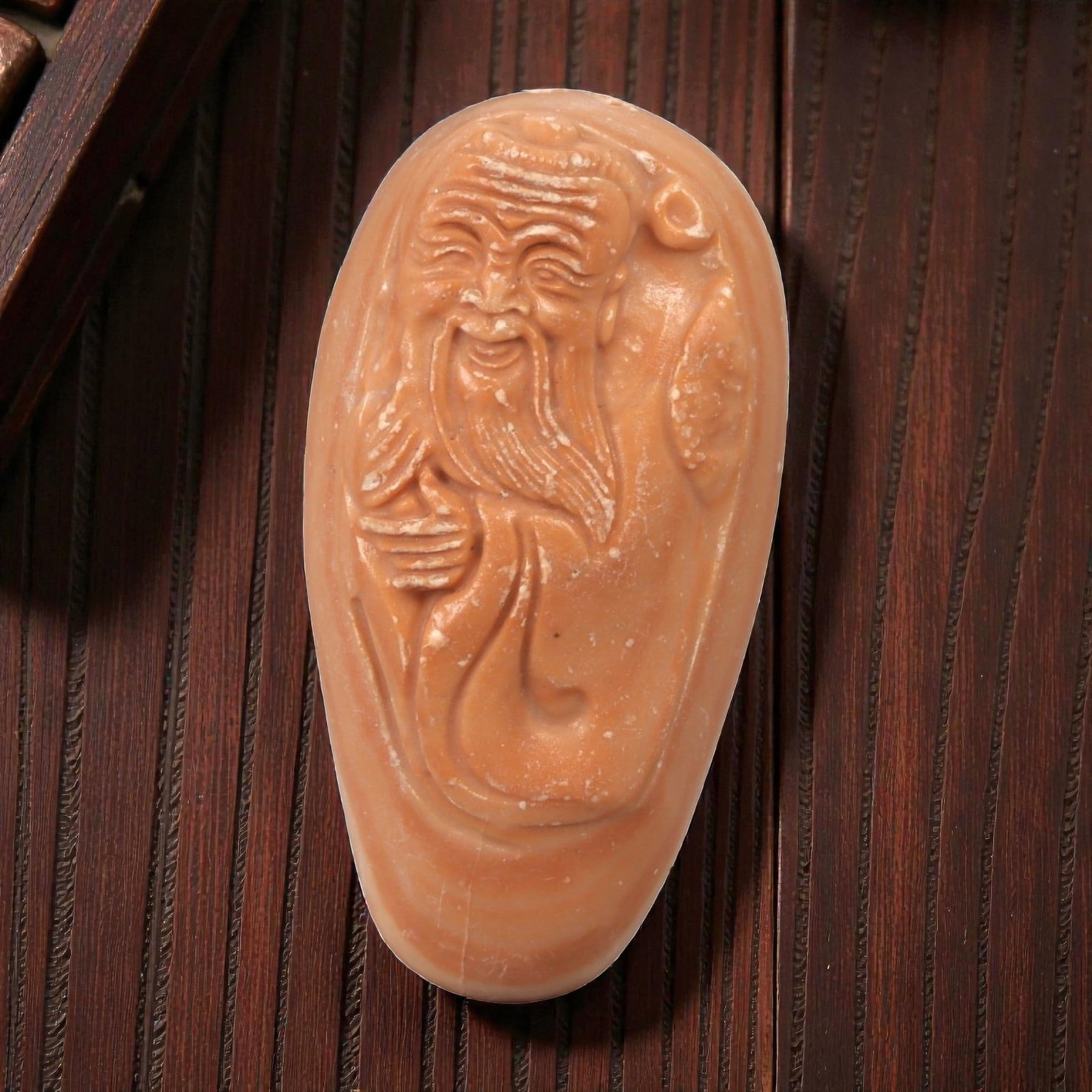 A Chinese Hardstone Figure of a Daoist Immortal, 23YB: Carved from reddish-brown hardstone with areas of lighter mottling and scattered surface inclusions, the object is worked as a vertical, elongated figure in relief. The figure is depicted with a long 