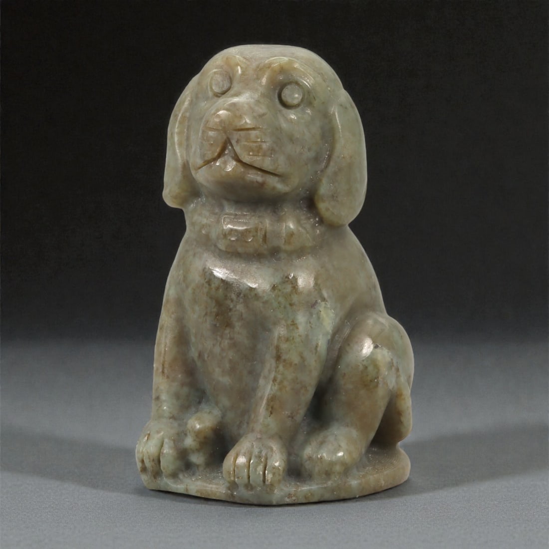 A Chinese Jade Seated Dog Figure, 20th Century, 89YA: Carved from grey-green jade with mottled brown inclusions and areas of soft translucency, the figure is modeled fully in the round as a seated dog shown upright on its haunches. The head is slightly r