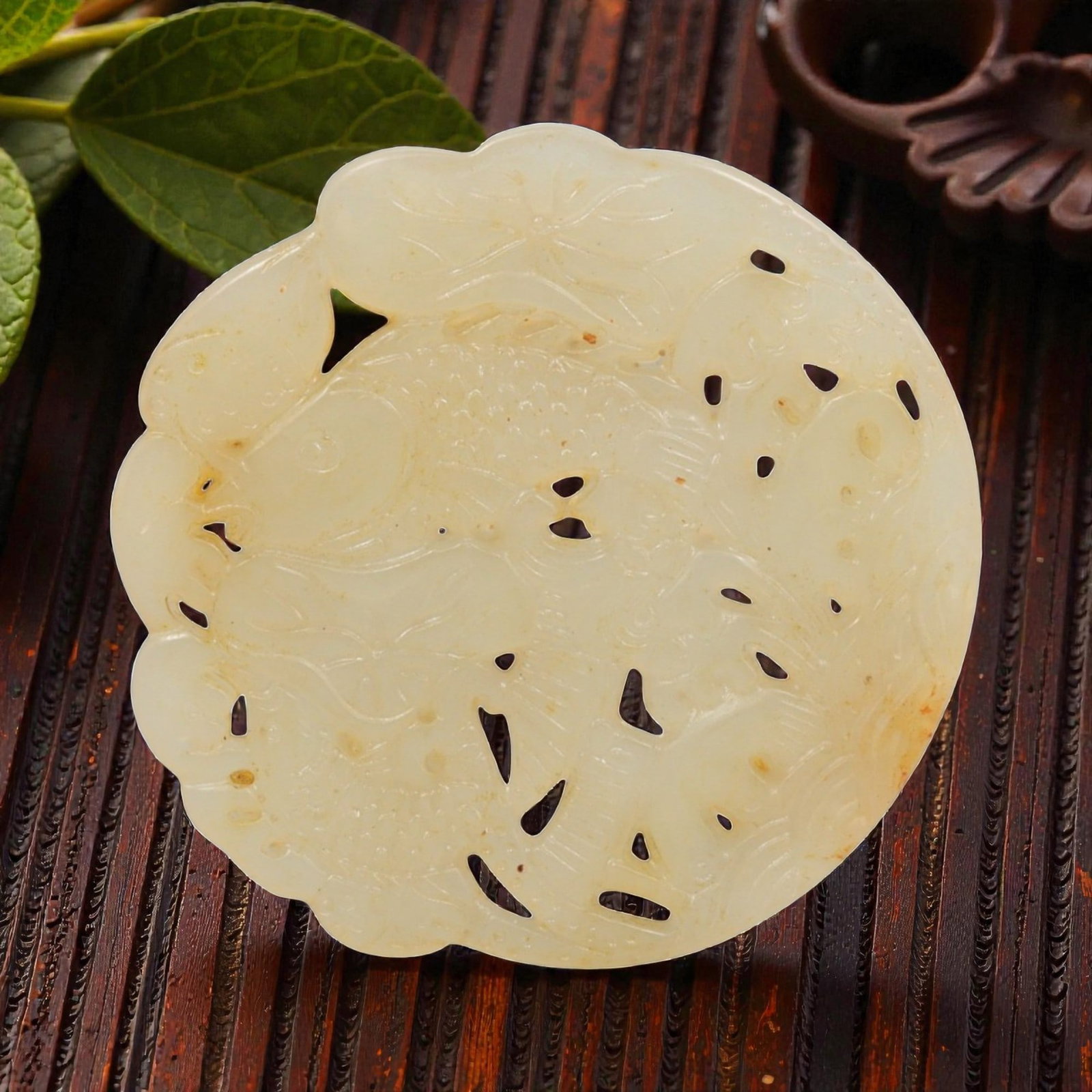 A Chinese Hetian Nephrite Jade Openwork Plaque Pendant, 34YC: Carved from pale celadon-toned nephrite jade with areas of light russet inclusions and natural mottling, the plaque is worked in openwork relief with a circular form and scalloped outer edge. The desi