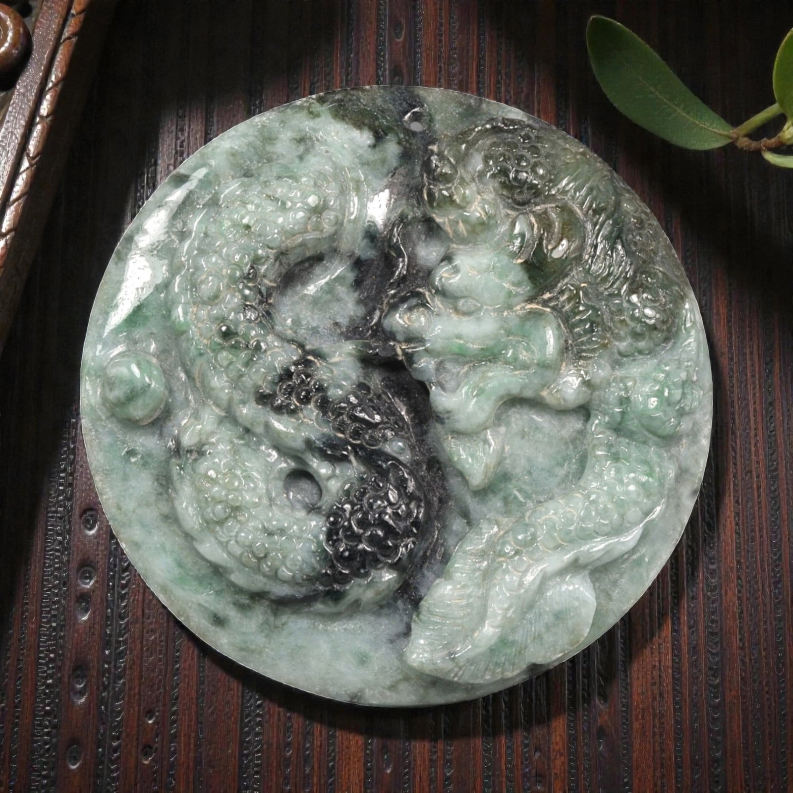 Natural Burmese Type A Jadeite Jade Carved Roundel Pendant, 4ZB: Carved from mottled green and dark-toned jade, the roundel is worked in high relief with a sinuous dragon shown in profile encircling a central aperture. The surface displays graduated scaling, a ridg