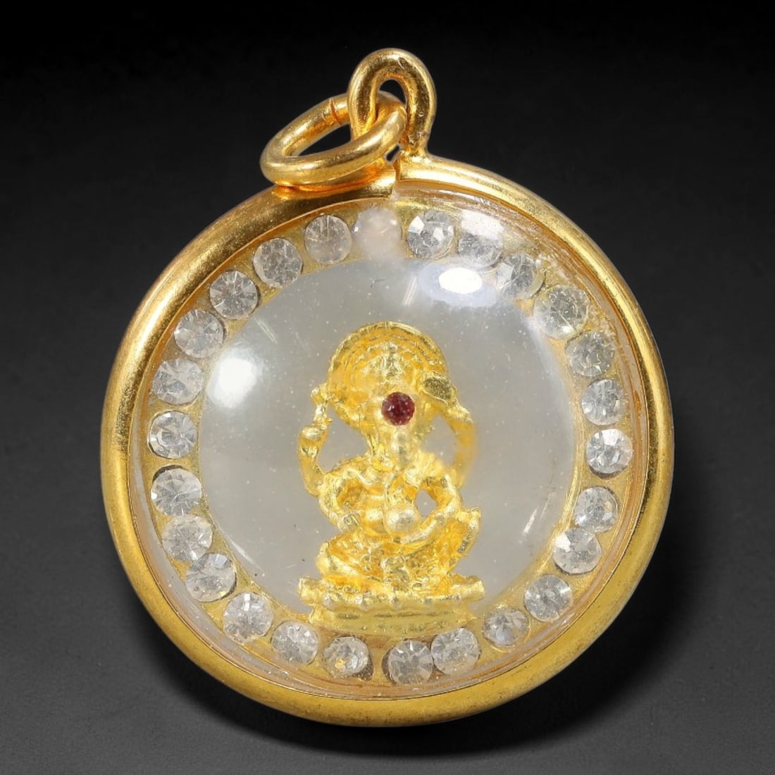 Thai Gold Plated Ganesha Amulet Pendant With Rhinestone Border, 82YY: Round gold plated casing with clear protective dome cover, sized 2.7x2.3cm. Front features a detailed miniature gold-toned seated Ganesha statue adorned with a small red gem accent at the forehead, su