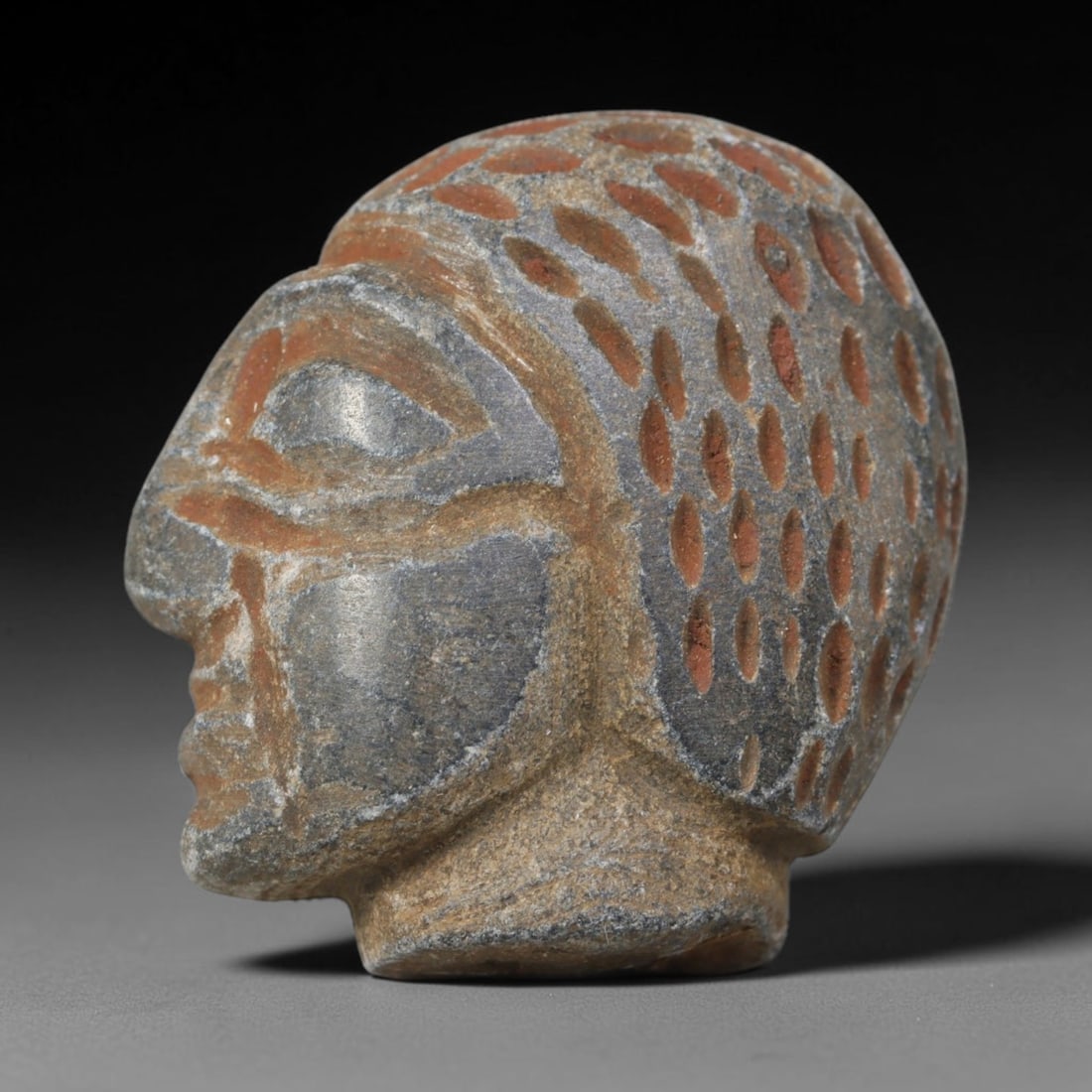 Antique Islamic Carved Stone Head, 35WT: Carved from grey stone with reddish pigment in the incised details, this artifact depicts a stylized human head in profile. The figure features simplified facial features, with a headdress or hairstyl