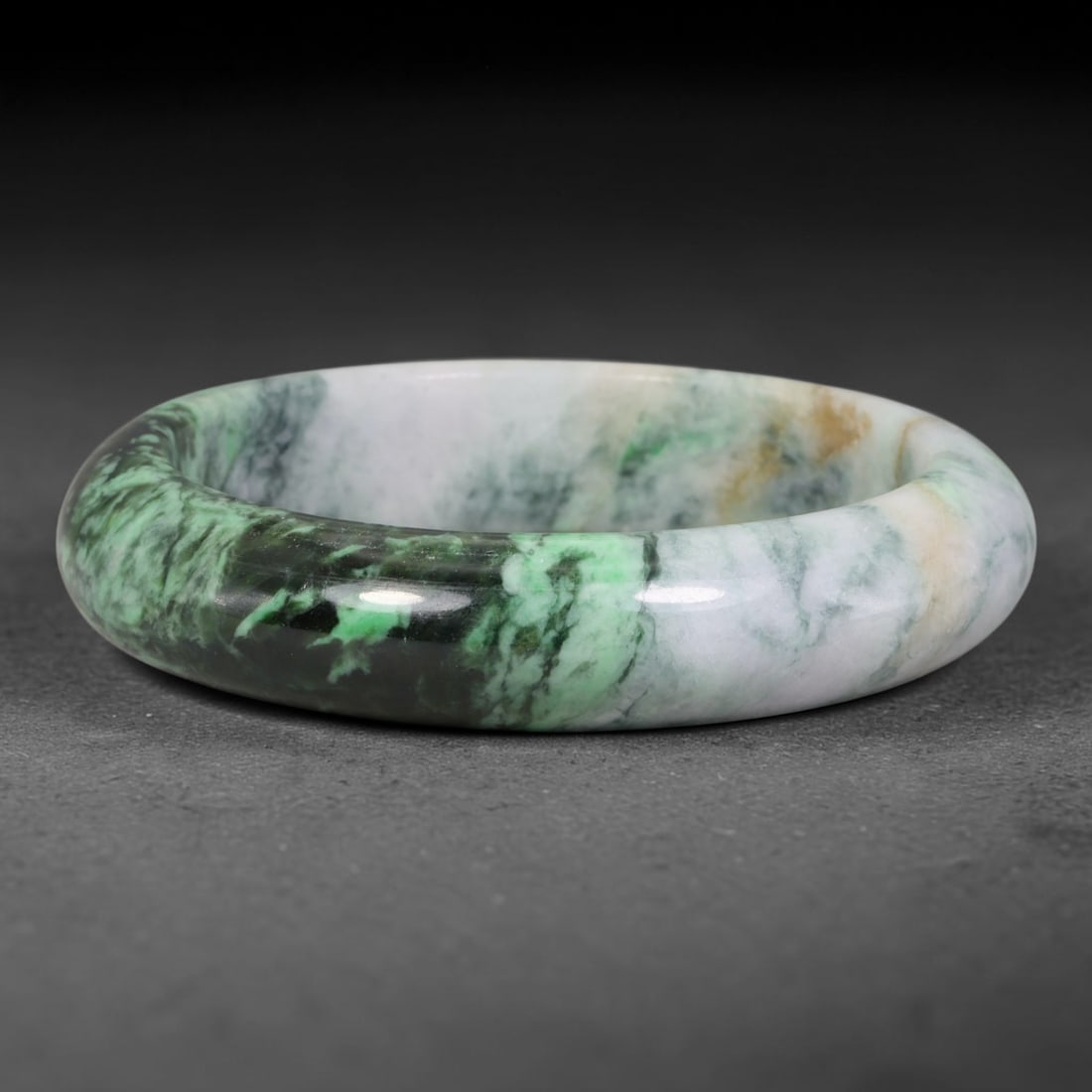 Natural Burmese Type A Variegated Floating Green Jadeite Jade Bangle Bracelet, 68TD: Polished from natural Burmese jadeite, this solid bangle exhibits variegated patterning with floating green veining set against pale white matrix, accented with subtle tan inclusions. Formed as a trad