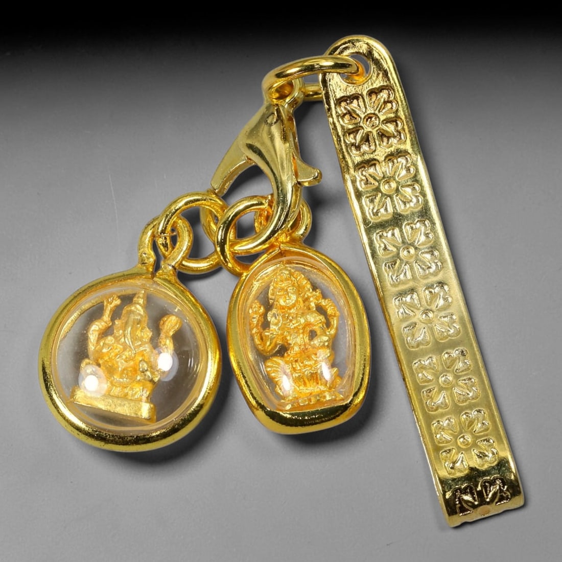 Dual Thai Amulet Charms Set with Floral Engraved Gold Clip, 24YY: Two mini golden deity pendants paired with a floral embossed gold clip, connected by lobster claw clasp and gold jump rings. One round clear cased amulet holds a detailed Ganesha figurine, while the o