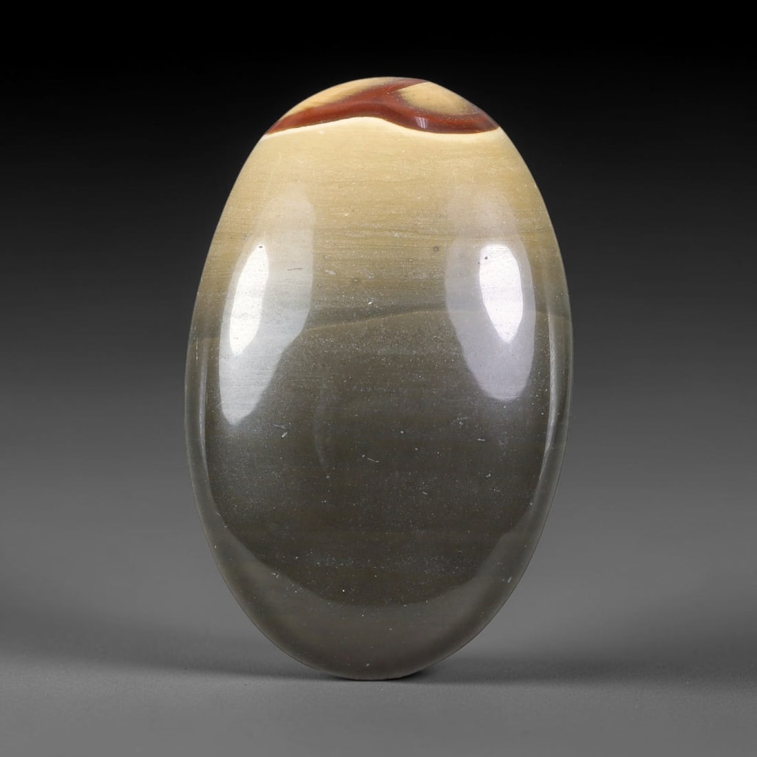 85.44ct Natural Polychrome Jasper Oval Polished Cabochon, 44TH: This polished specimen is natural polychrome jasper, shaped as a smooth oval cabochon. It features subtle horizontal banding transitioning from pale beige and cream through grey tones, accented by a t
